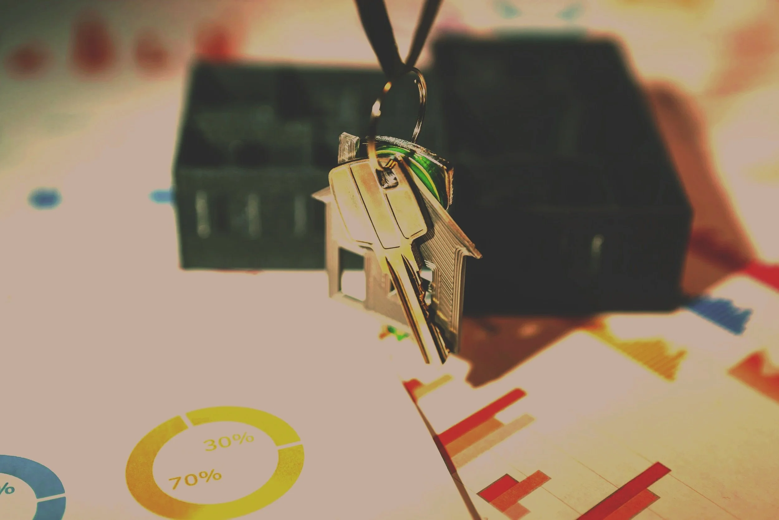 Close-up view of a house-shaped keychain hanging from a black keyring, with various colorful charts and graphs on paper underneath, and a blurred black organizer in the background.