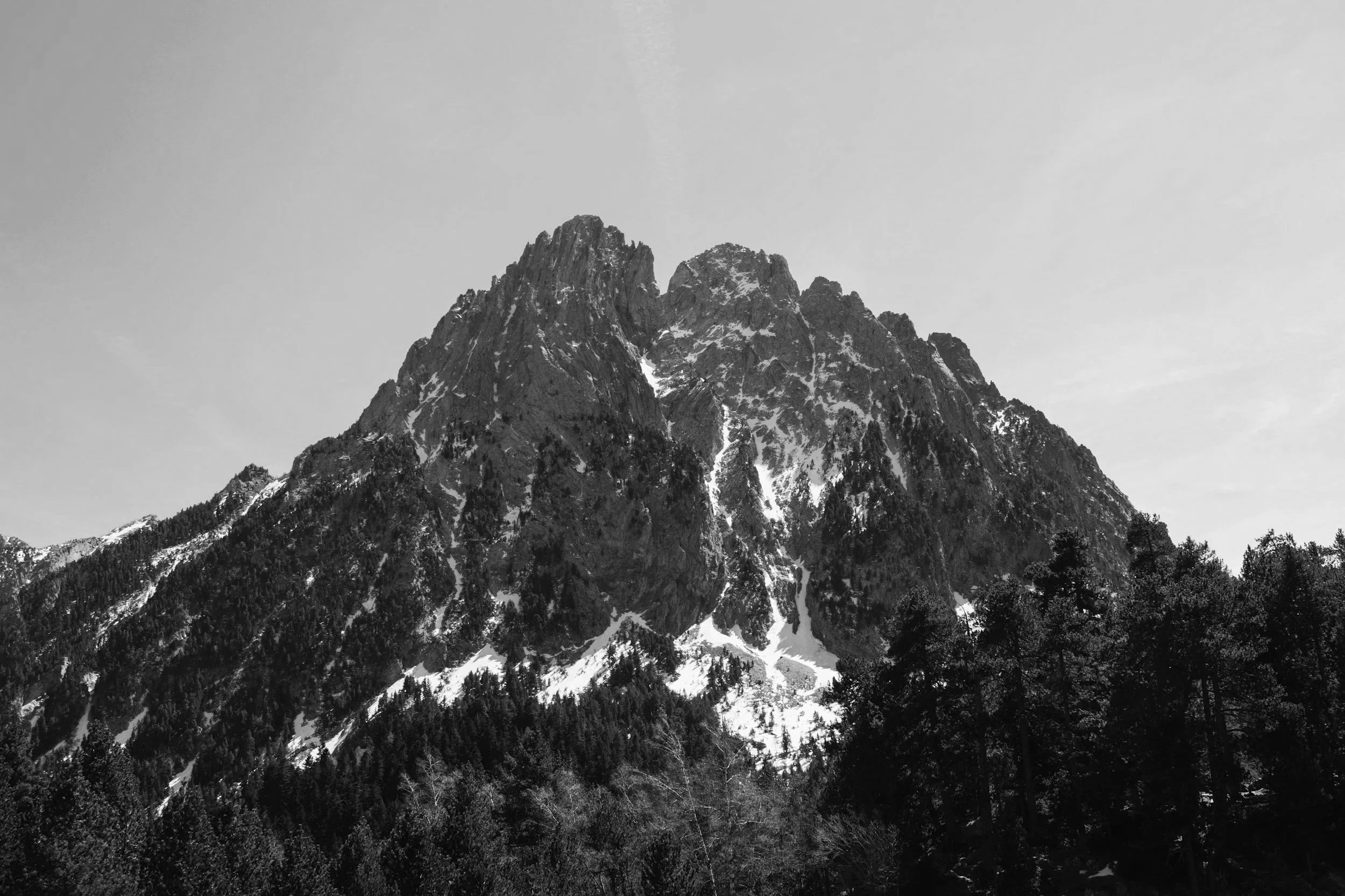 Black and white photograph of a tall, rugged mountain with some snow patches, surrounded by trees at its base and a clear sky above.