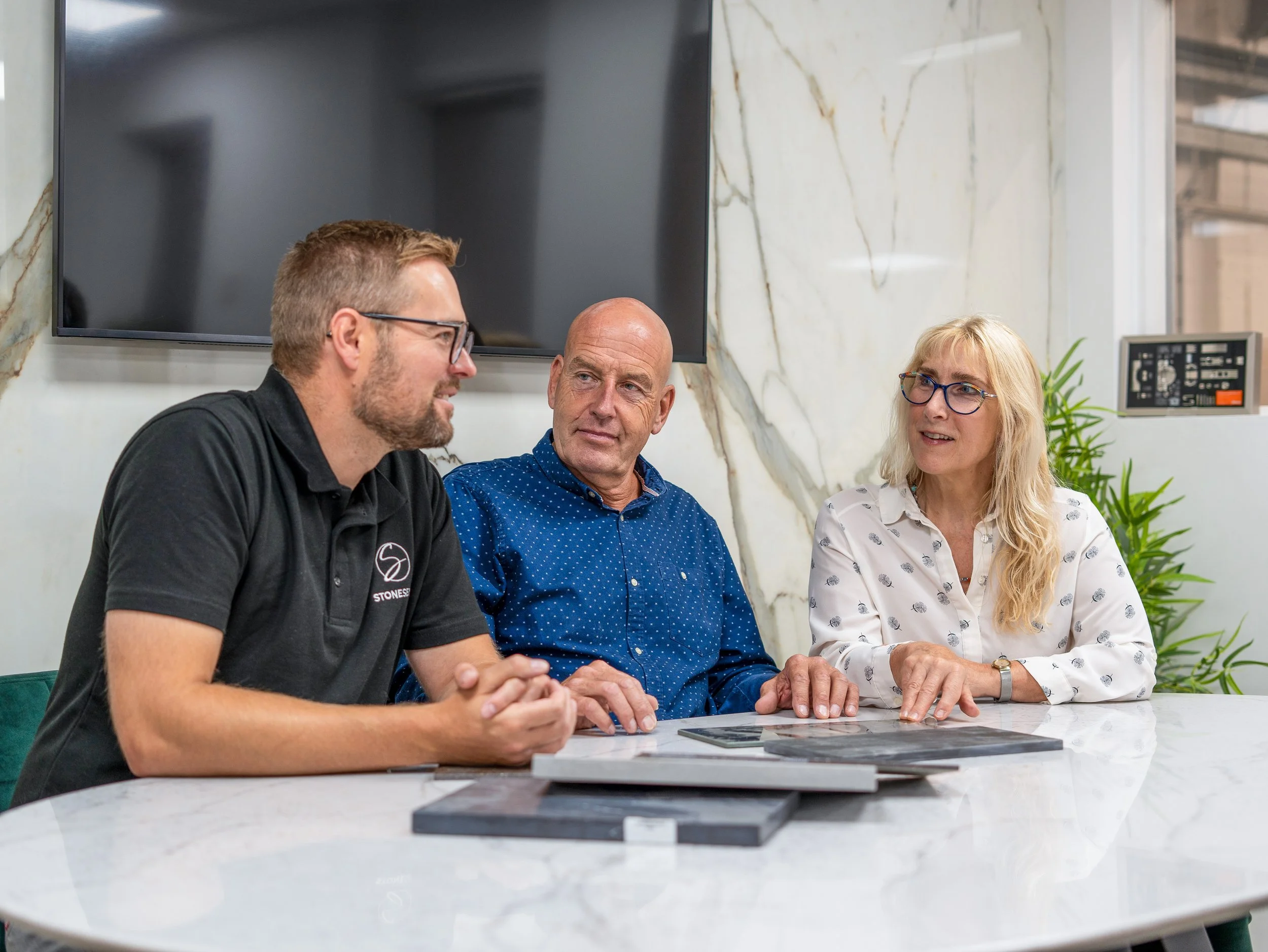 Three people sit at a marble table in a modern office, engaged in a discussion. The man on the left wears glasses and a black polo shirt, the man in the middle has a shaved head and a blue polka-dot shirt, and the woman on the right has blonde hair, glasses, and a white blouse with small patterns. They are surrounded by a large black screen and some documents or samples on the table.