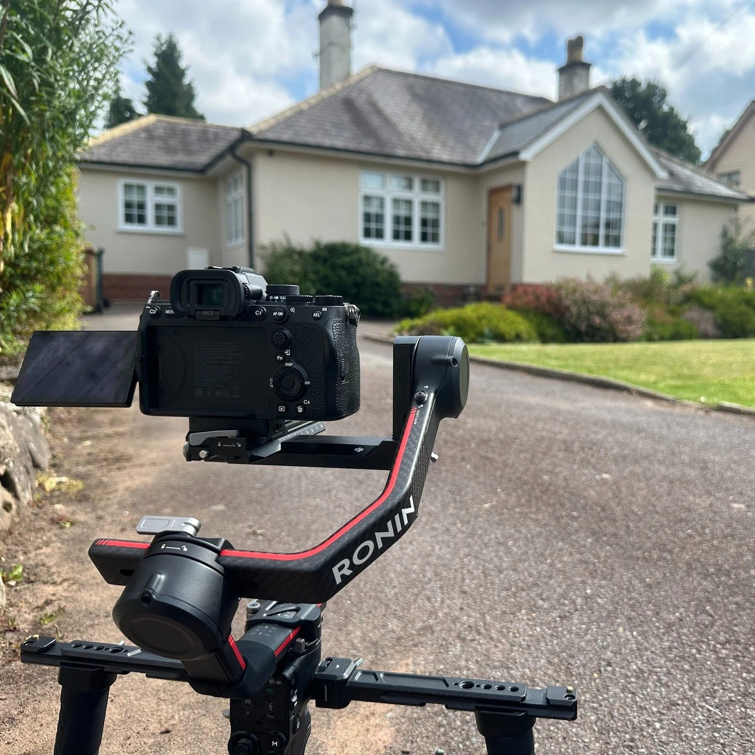 A camera mounted on a gimbal stabilizer set up on a paved driveway in front of a house, with the house featuring a large window and a small garden in the background.