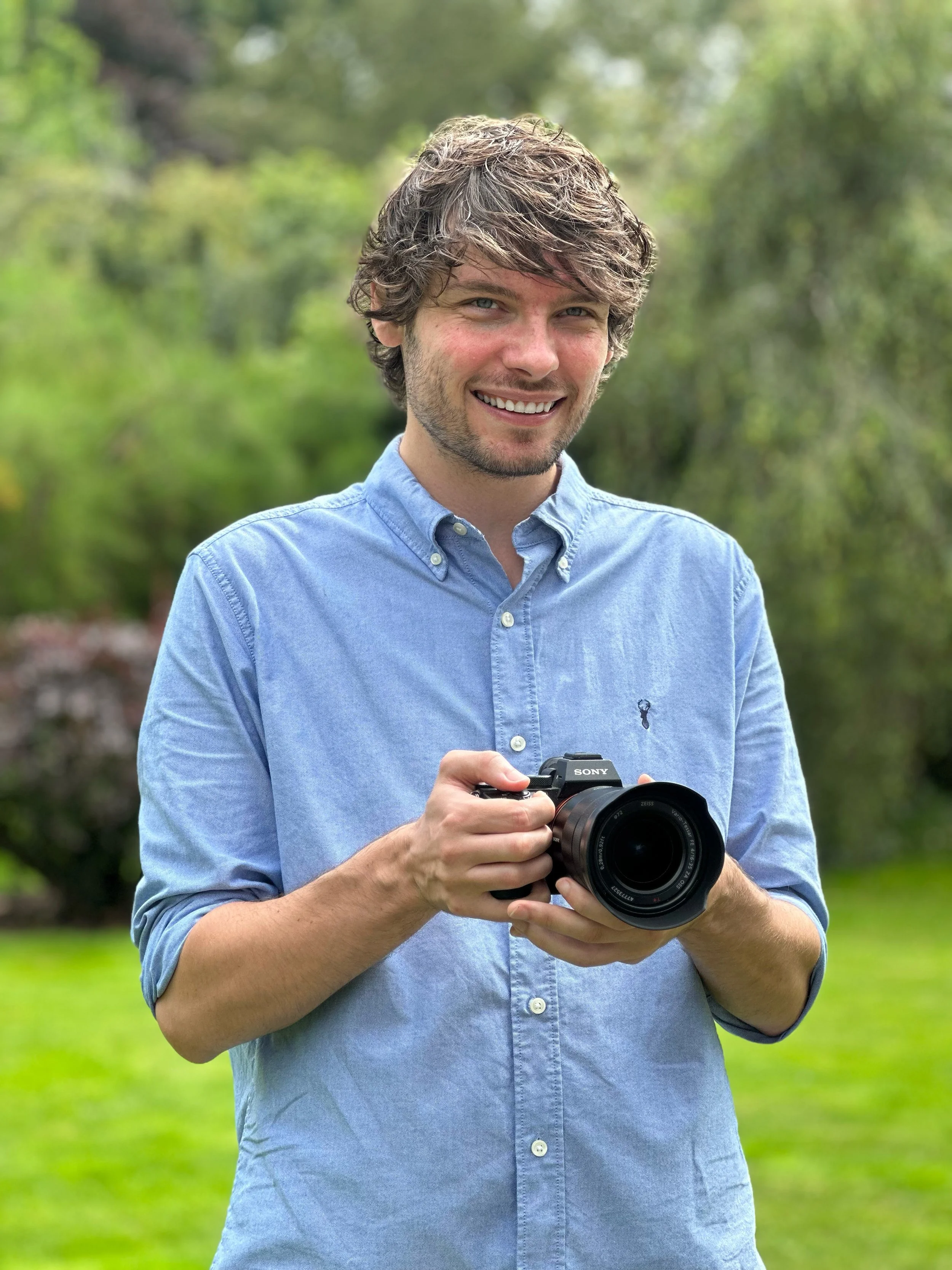 A smiling man in a light blue button-down shirt holding a Sony camera outdoors in a park with green trees and grass.