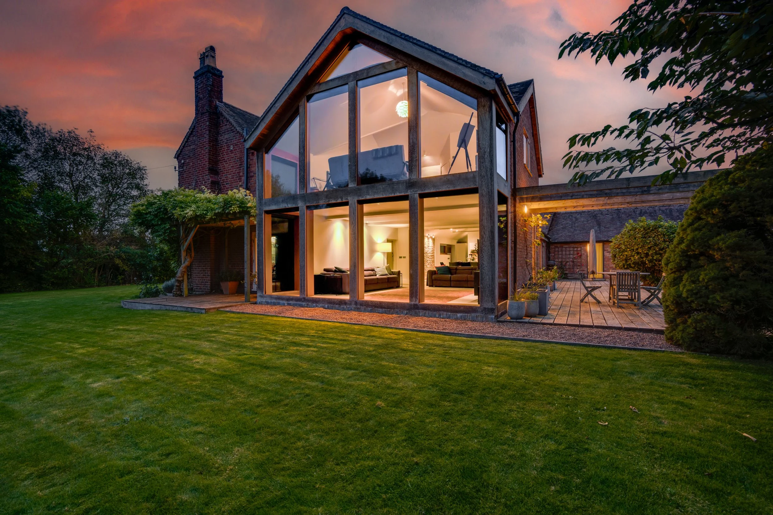 A house with a brick exterior and large windows showing a lit interior at dusk, surrounded by a green lawn and trees.