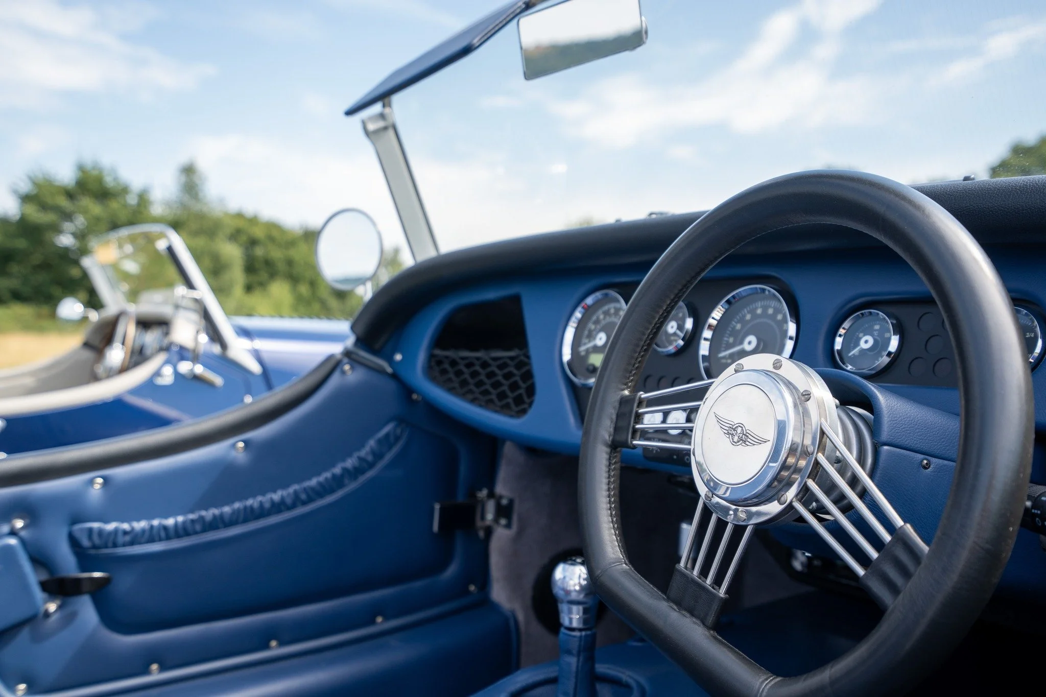 Inside view of a vintage blue race car with a leather steering wheel that has a winged emblem at the center, and a dashboard with multiple round gauges, with a reflection of trees and sky seen through the windshield.