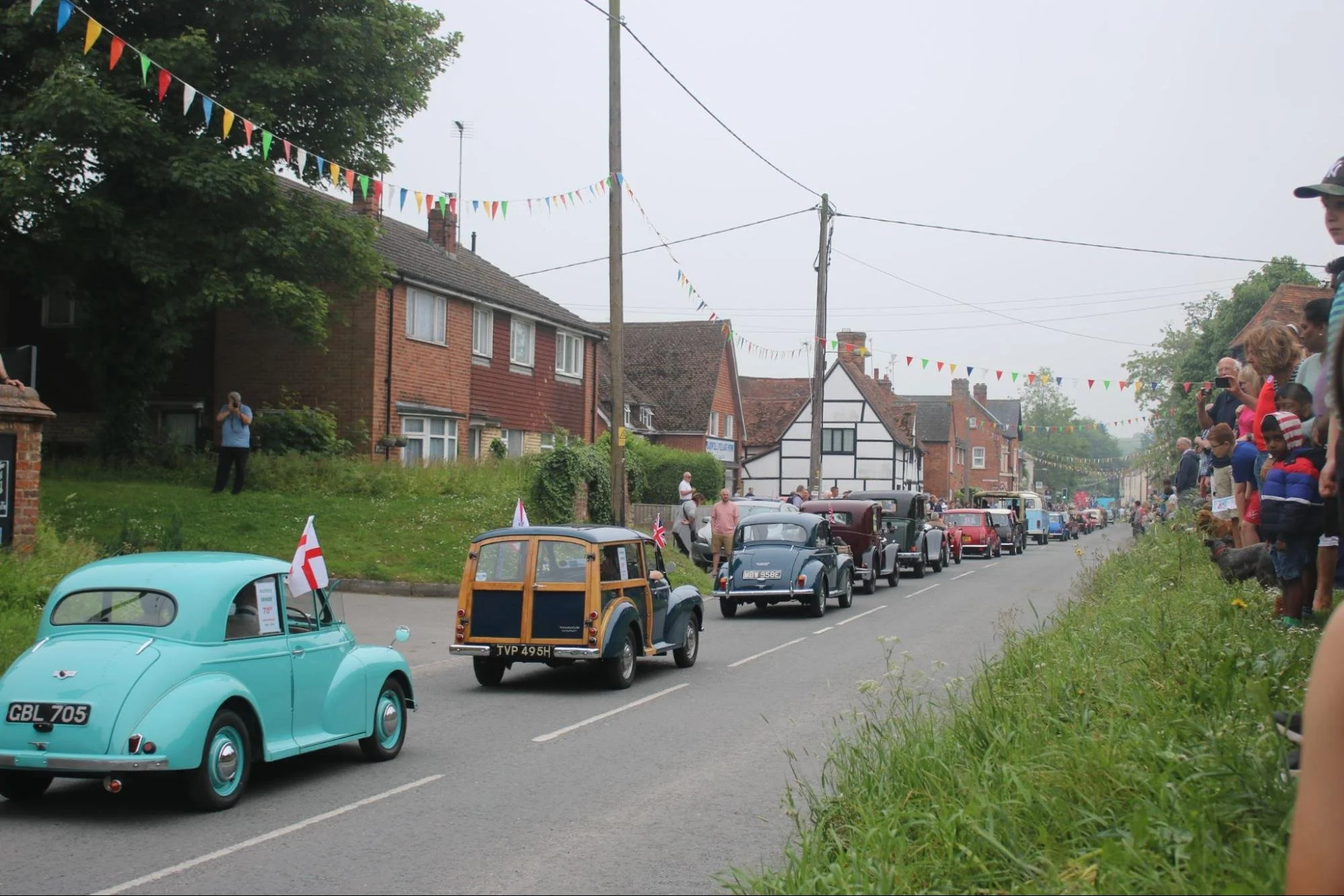 A parade of vintage cars driving down a suburban street decorated with colorful pennant banners, with spectators watching from the side of the road.