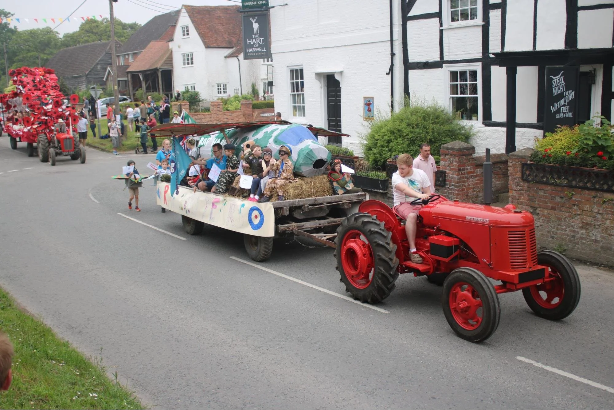 A parade float designed as a World War II-themed plane with children dressed as soldiers riding on it, being pulled by a vintage red tractor.
