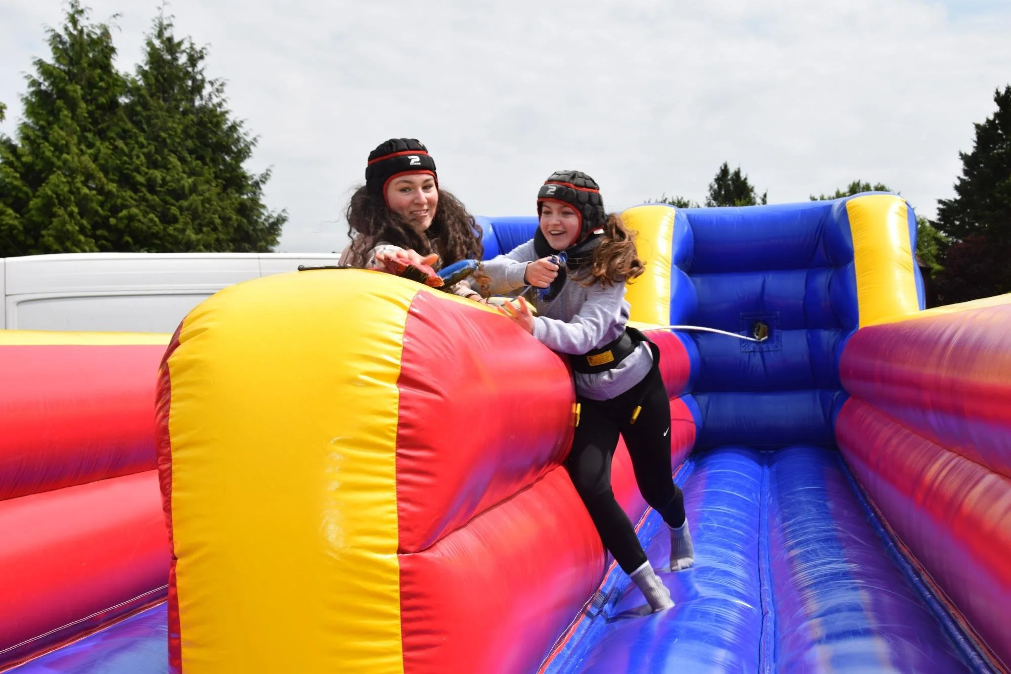 Two young girls wearing helmets enjoying a ride on an inflatable obstacle course outdoors with trees and cloudy sky in the background.