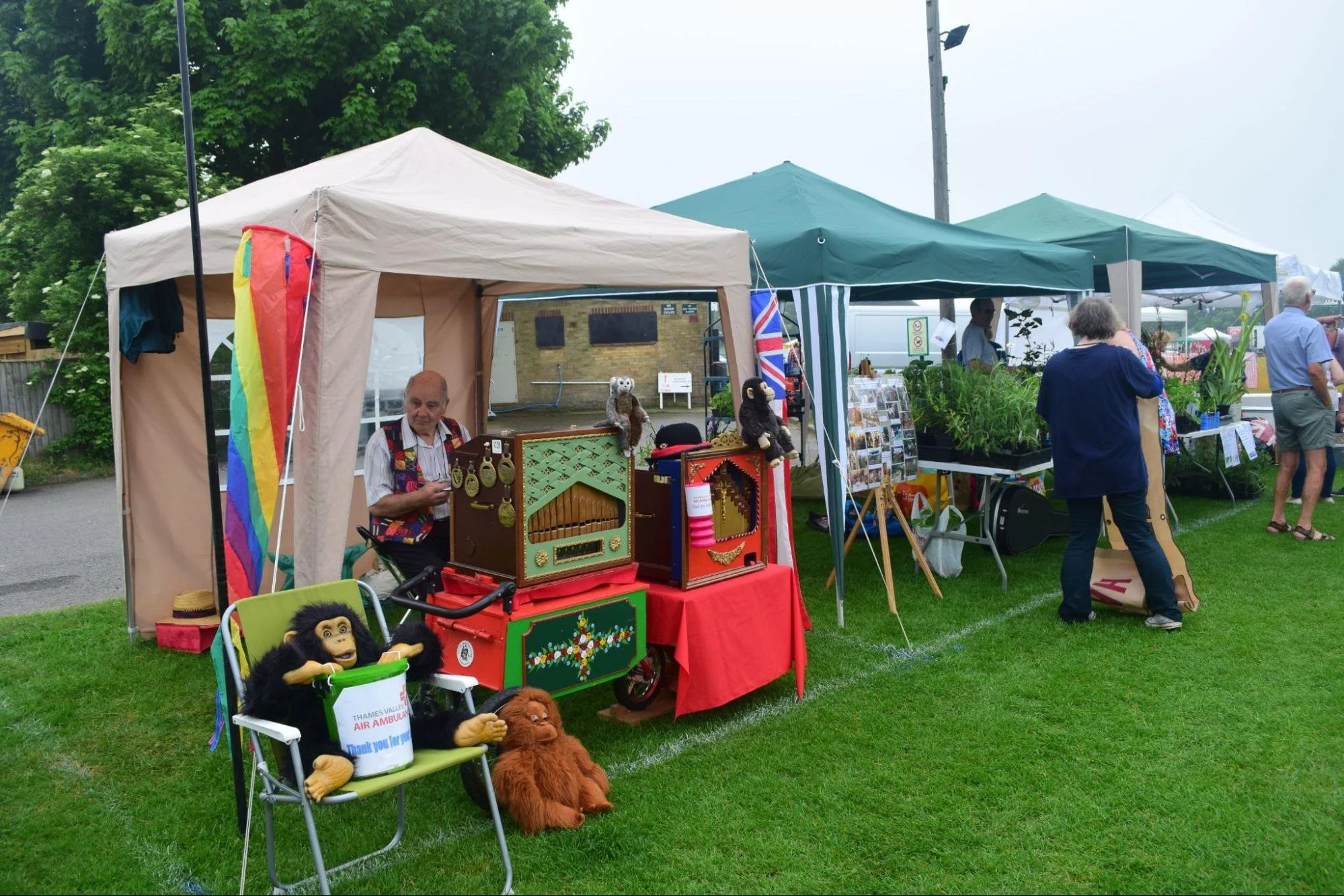 Outdoor market stall with vintage toys, puppets, and stuffed animals, beside tents selling plants and other items, with people browsing and green trees in the background.