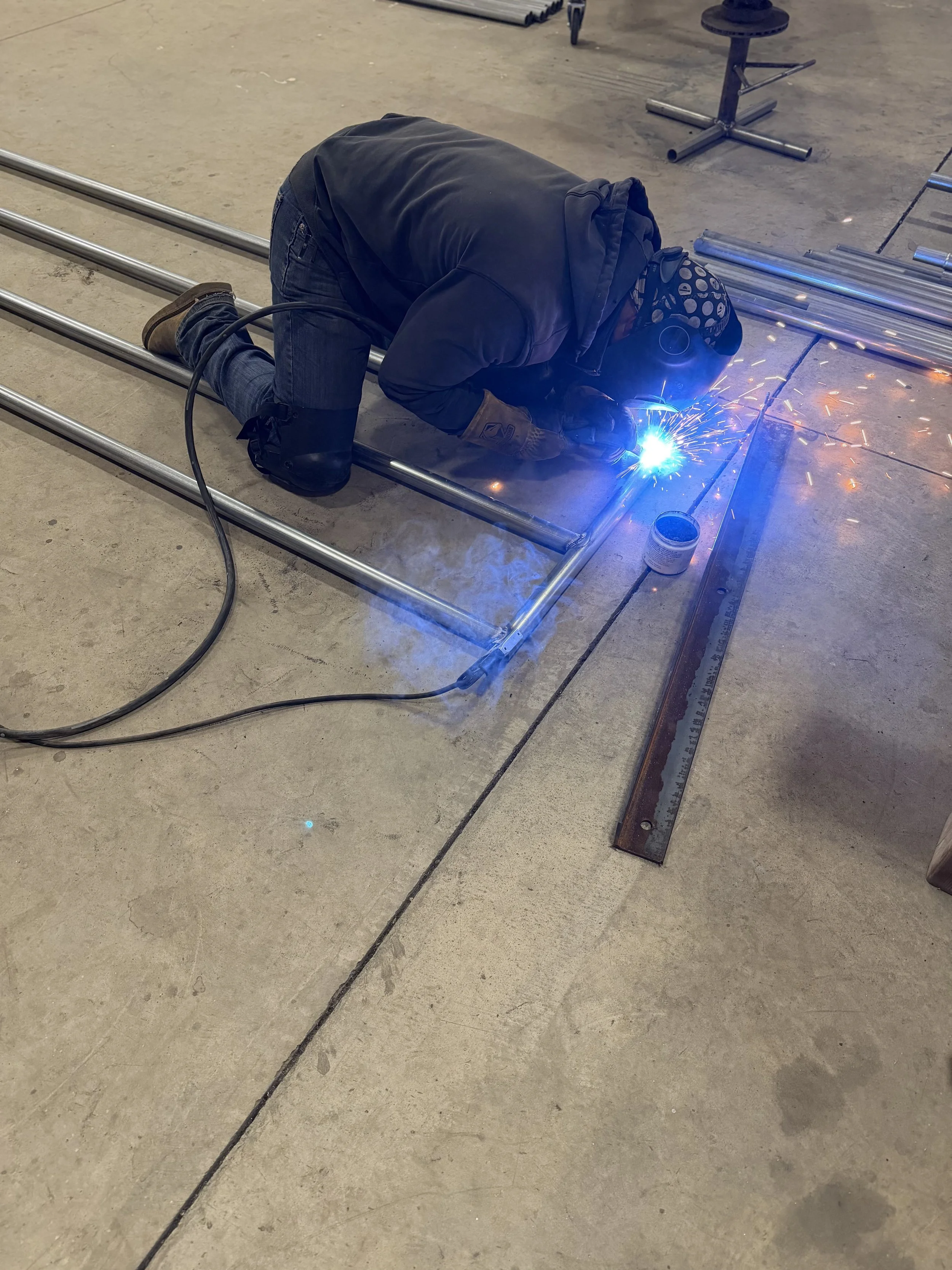 A person wearing protective gear welding metal pipes on a concrete floor, sparks flying from the weld.