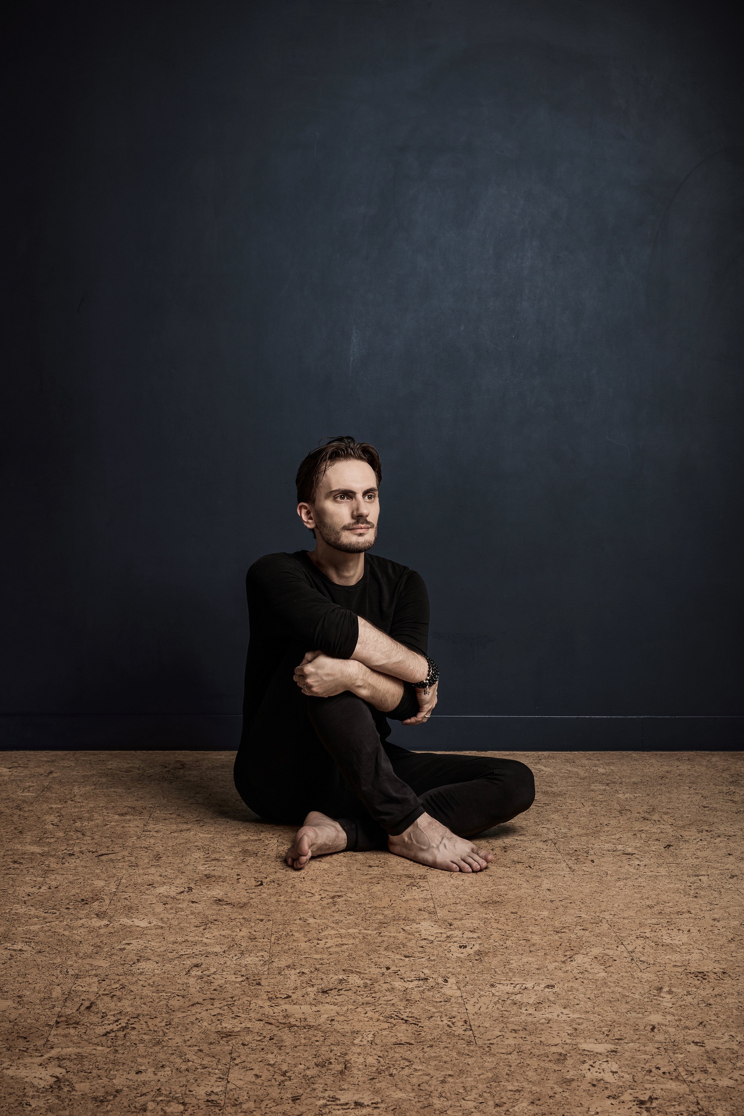 Un homme assis par terre, les bras croisés autour de ses jambes, regardant vers la droite, avec un mur foncé en arrière-plan.
