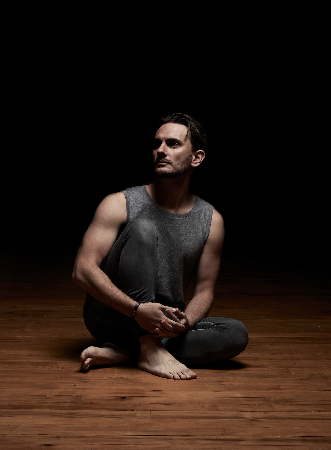 Homme assis sur un sol en bois, habillé d'un débardeur gris, dans un environnement sombre. Alex Blake Yoga
