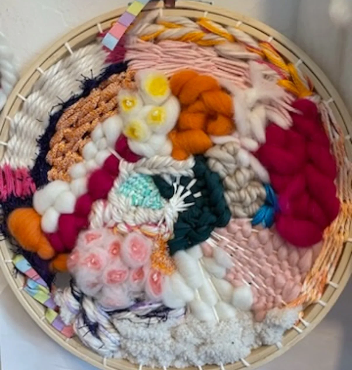A round woven basket filled with various colorful yarns and fibers, including wool, cotton, and roving, arranged in sections.