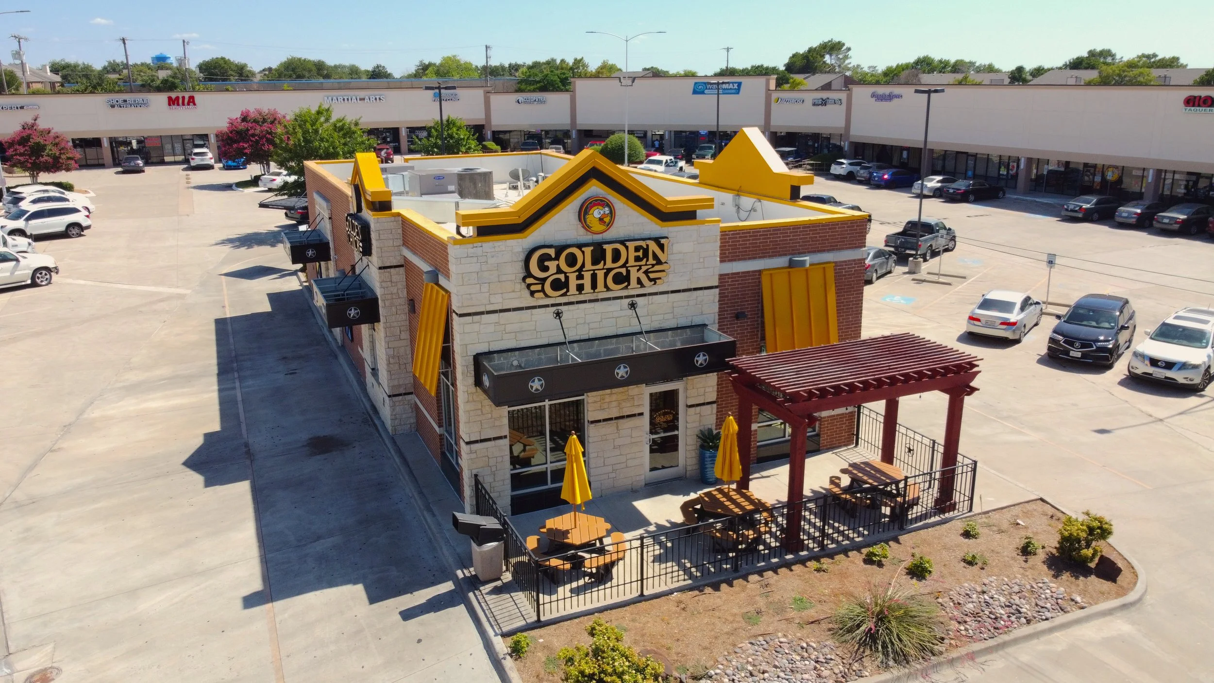 A Golden Chicken restaurant building with outdoor seating area featuring yellow umbrellas, a red pergola, and surrounding landscaped plants, parking lot in background
