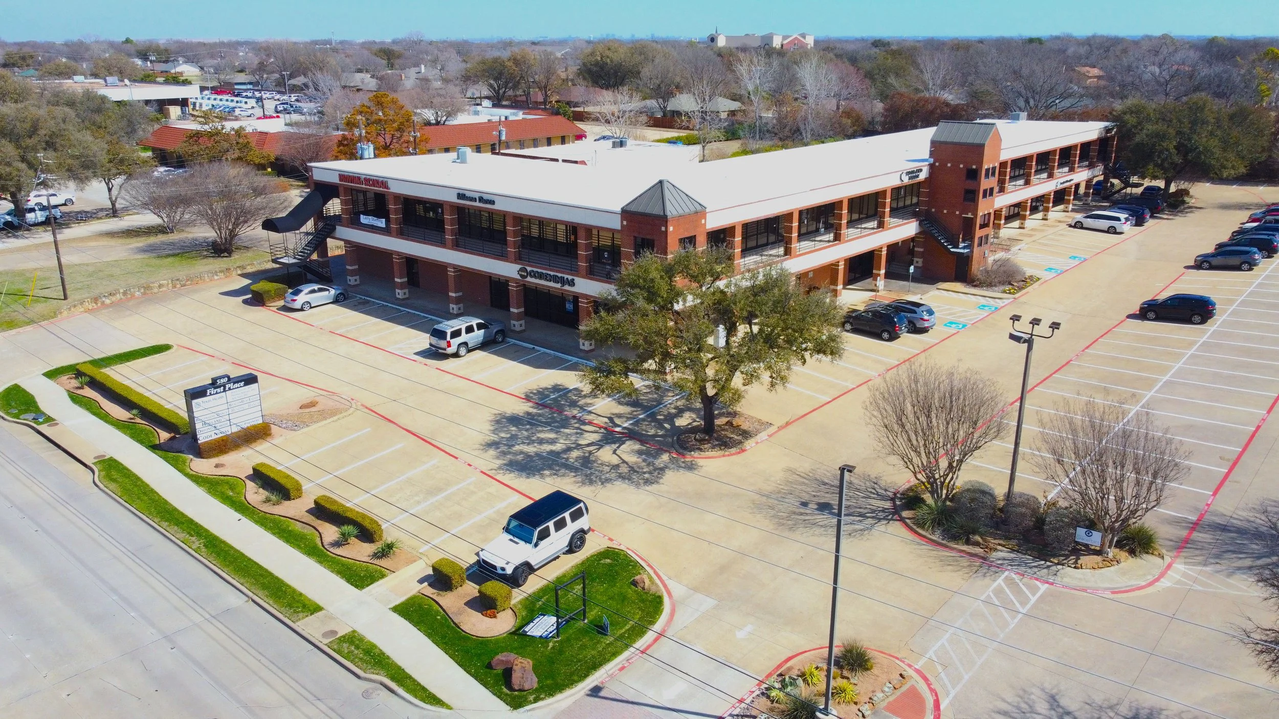 Aerial view of a strip mall with parking lot in front, featuring cars parked near a two-story brick building labeled 'First Place.' Surrounding the building are trees, landscaping, and street lamps, with other buildings and trees visible in the background.