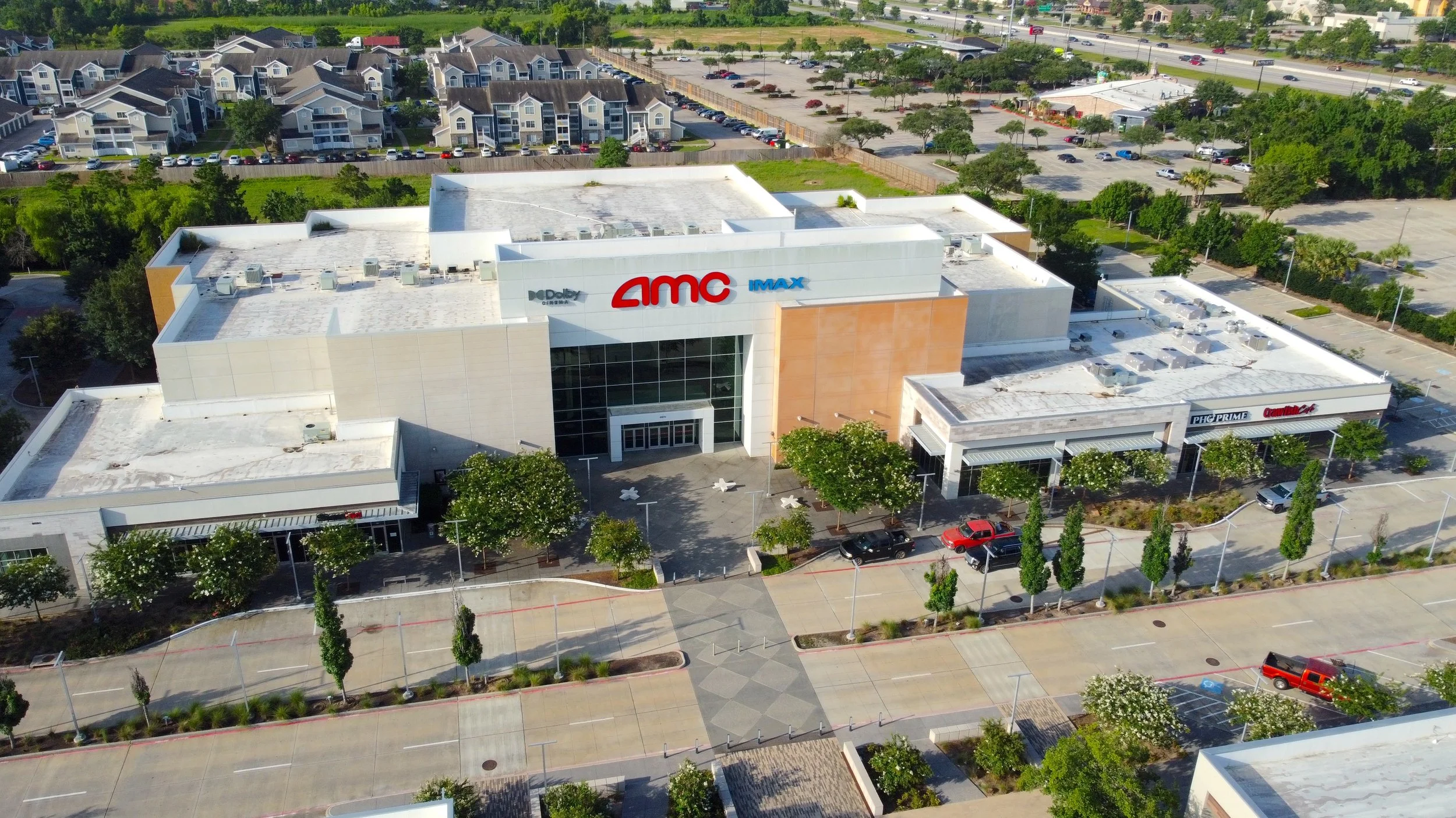 Aerial view of a large shopping mall with parking lot, featuring the AMC theater with prominent red and blue signs, surrounded by trees and other commercial buildings.
