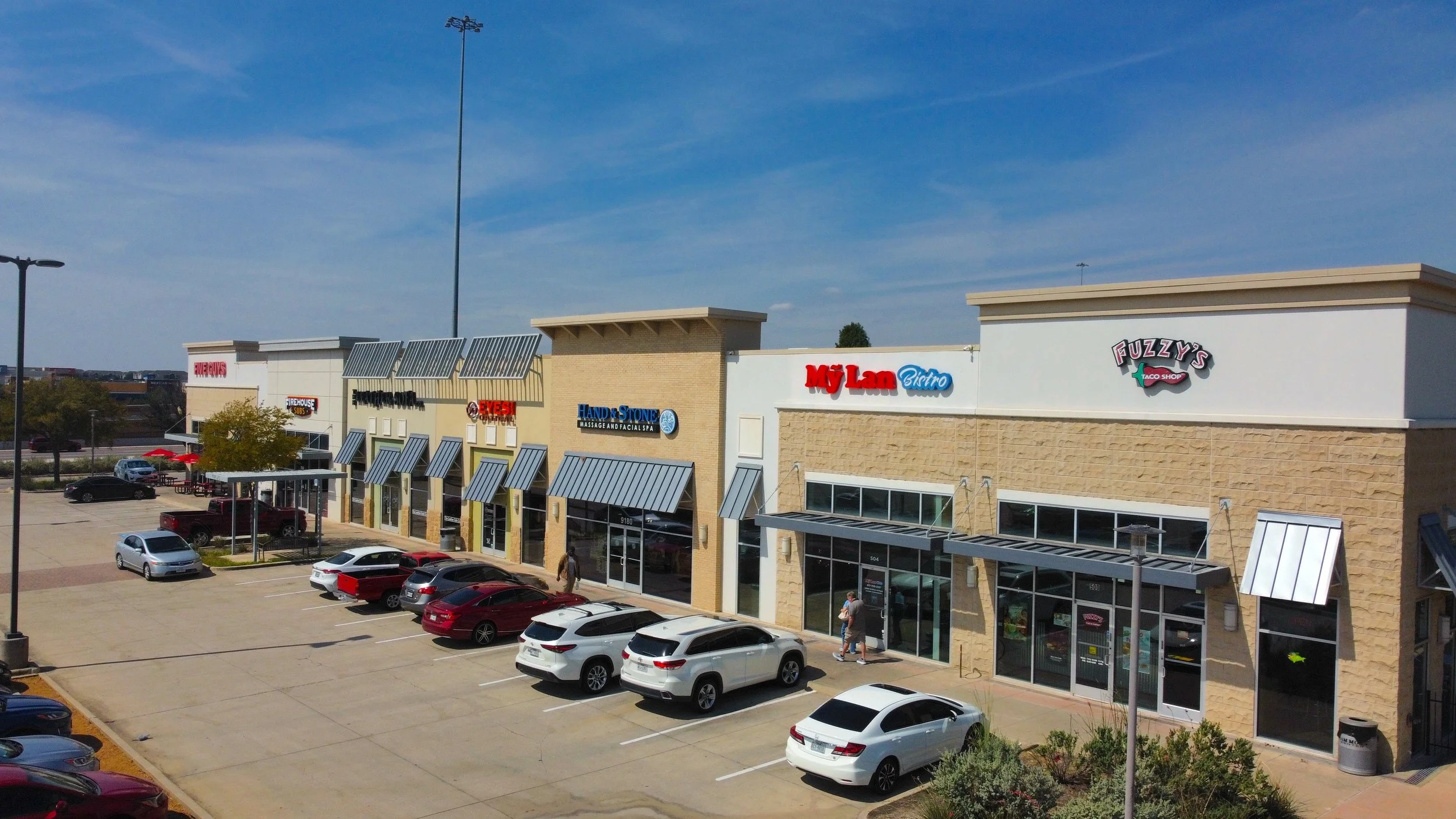A shopping plaza with various storefronts including MF Lan Bistro, Fuzzy's Taco Shop, Hand & Stone Massage and Facial Spa, and a fireworks store, with cars parked in front on a sunny day.