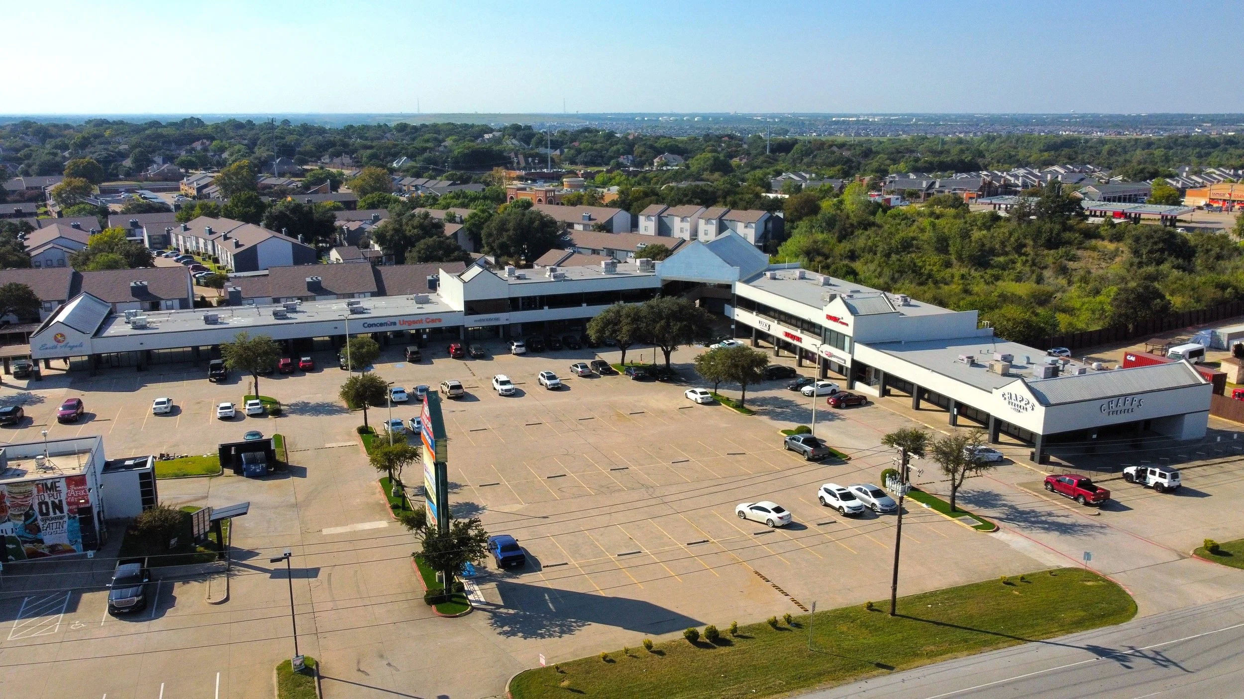 A large, mostly empty shopping center parking lot with a few parked cars, surrounding a retail strip mall with multiple storefronts, trees, and a signpost. In the background, residential neighborhoods and greenery extend to the horizon.