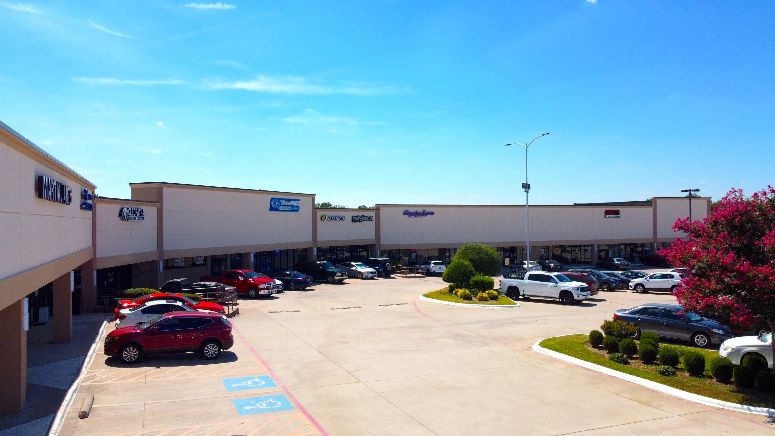 Shopping plaza parking lot with several parked cars, small landscaped islands, and storefronts under a blue sky.