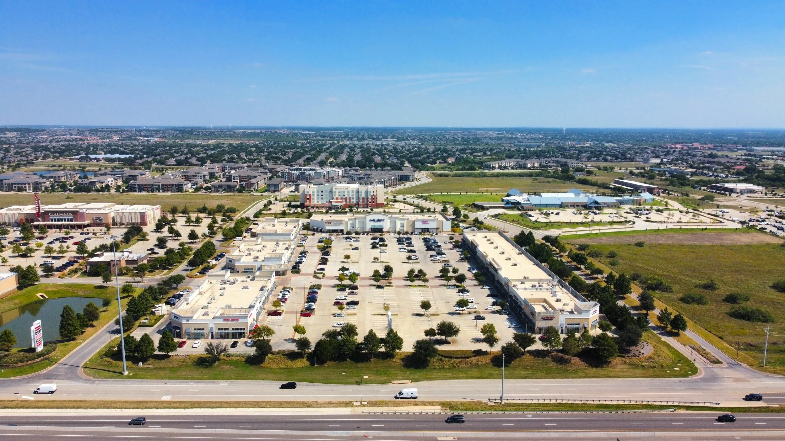 Aerial view of a shopping mall parking lot with multiple storefronts, surrounded by trees and roads, with a highway in the foreground.