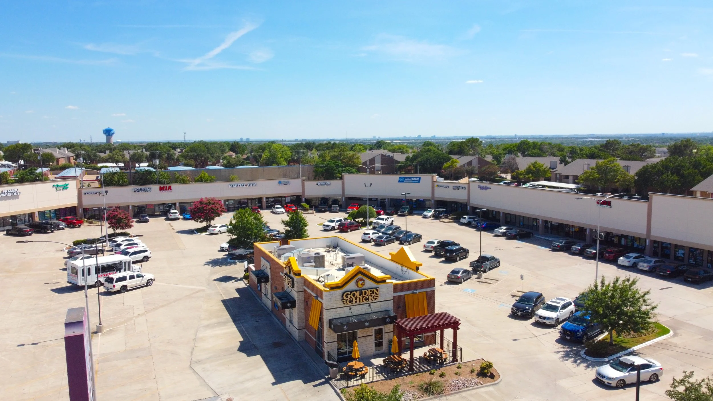 A shopping center parking lot with various parked cars, a Golden Chick restaurant in the foreground, and single-story retail stores in the background. There are trees and a clear blue sky.