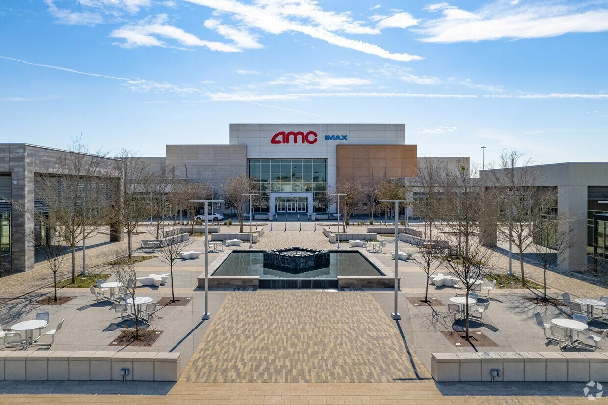 Empty outdoor shopping plaza with trees, benches, tables, and a fountain in front of an AMC IMAX theater building under a blue sky with clouds.