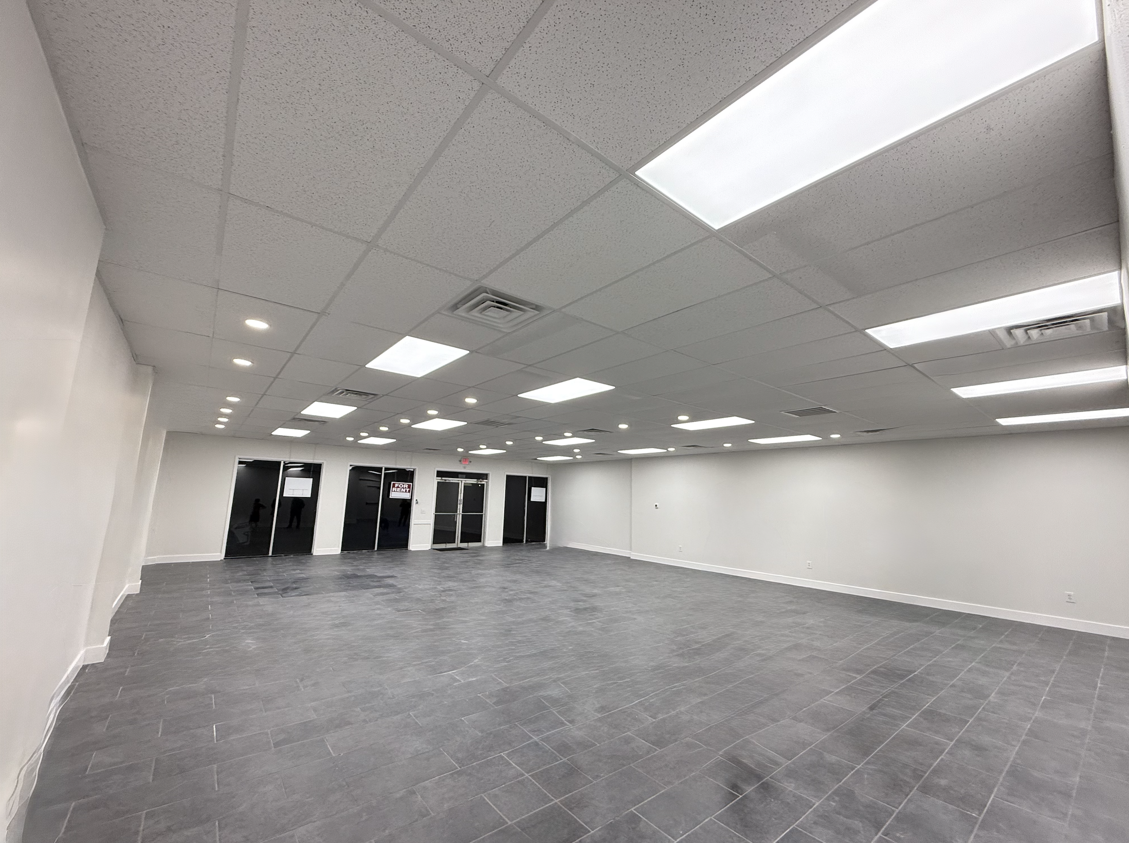 Empty commercial space with gray tiled floor, white walls, and dropped ceiling with fluorescent lights and vents. The before picture of a tenant representation showcase.