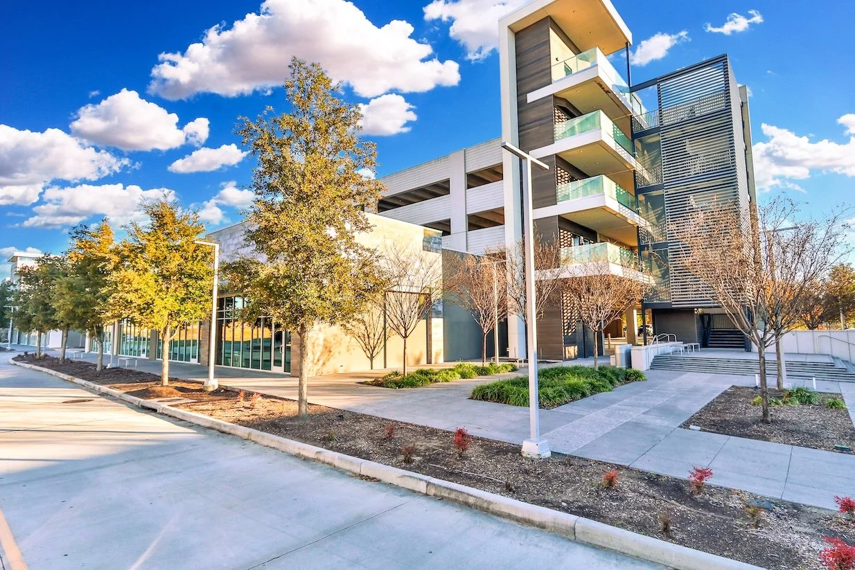 Modern multi-story building with glass balconies, surrounded by trees with some autumn foliage, under a partly cloudy blue sky.