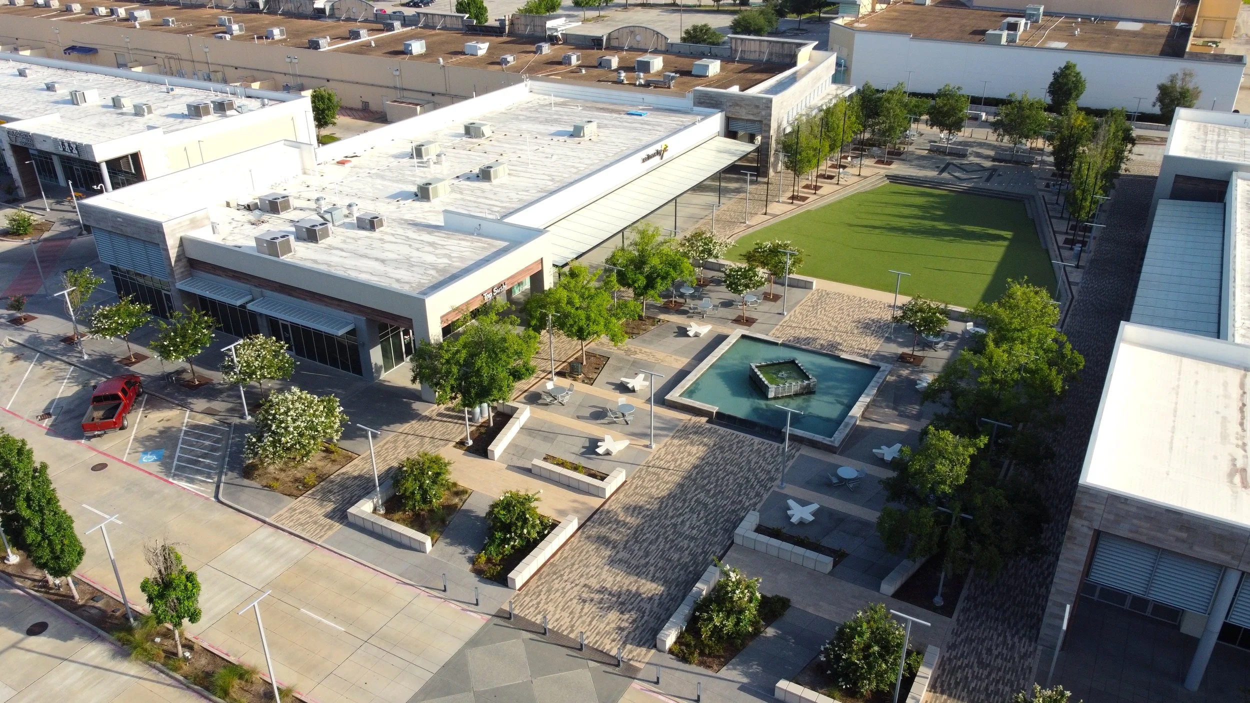 Aerial view of a modern shopping center with outdoor seating, a fountain, a parking lot, and a green lawn area.
