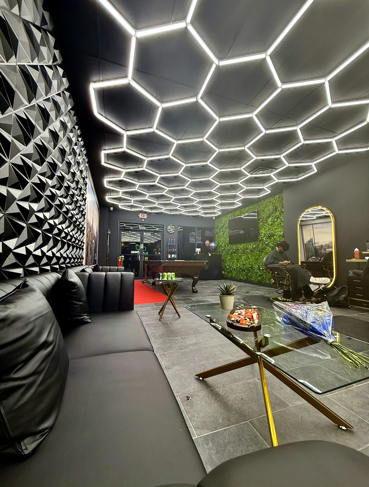 The after picture of a tenant representation showcase. Modern barbershop with black leather sofas, a glass coffee table with flowers, a potted plant, a pool table, a green wall, and geometric ceiling lighting.