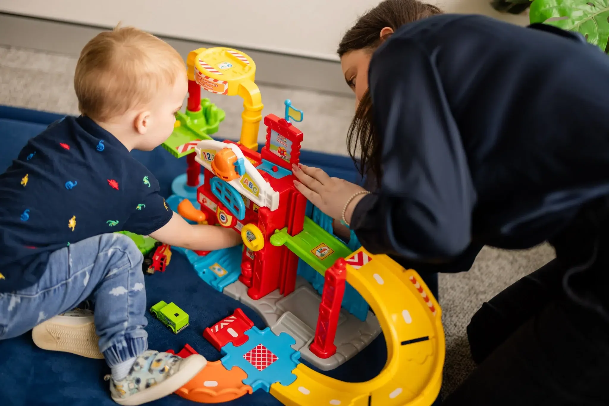 A young boy and a woman playing with a colorful toy race track set indoors. The boy is kneeling and reaching for the toys, while the woman looks on and helps.