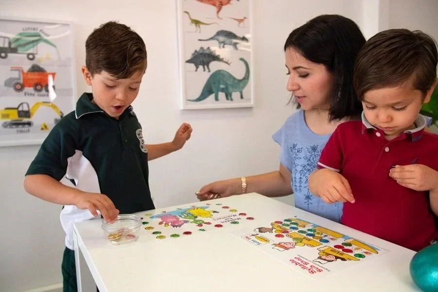A woman and two young boys playing a colorful board game on a white table in a room with dinosaur artwork on the wall.