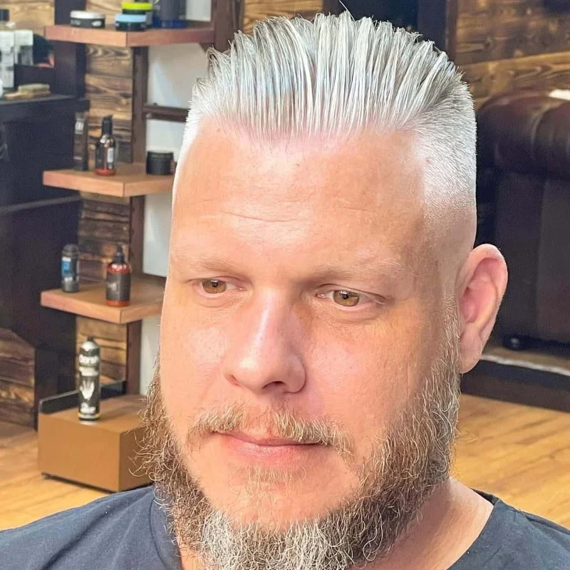 Man with a styled, platinum blonde comb-over hairstyle and a beard, sitting in a barbershop.