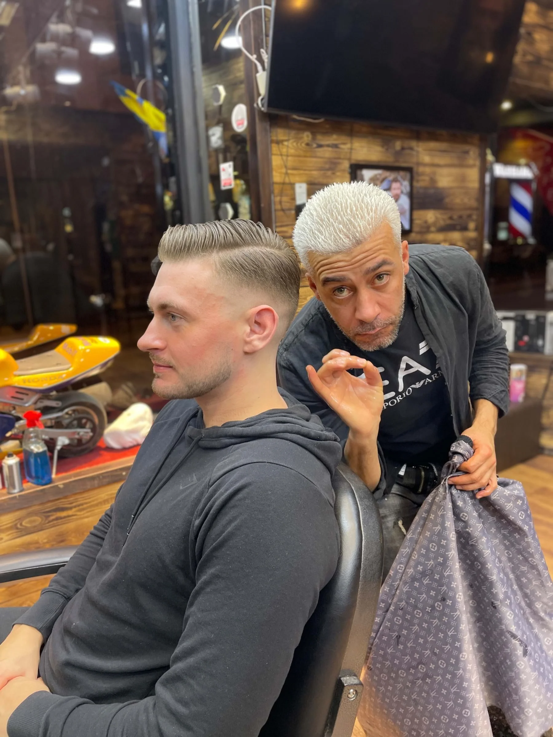A man with silver dyed hair getting a fresh haircut at a barbershop, seated in a chair. The barber, with platinum blonde hair, is holding a bag and making an OK sign with his hand while looking at the camera. The background shows wooden walls, a TV, and barber equipment.