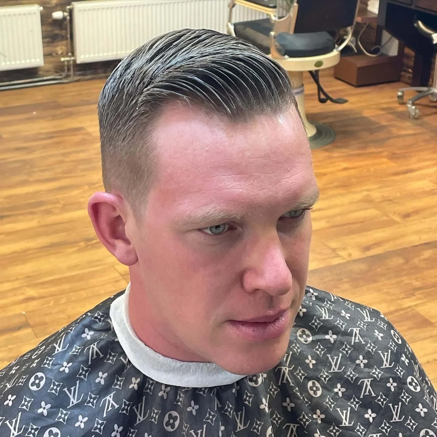 A man with a fresh, slicked-back haircut sitting in a barbershop. He is wearing a black barber cape with Louis Vuitton monogram pattern, and the background shows wooden flooring and barber chairs.
