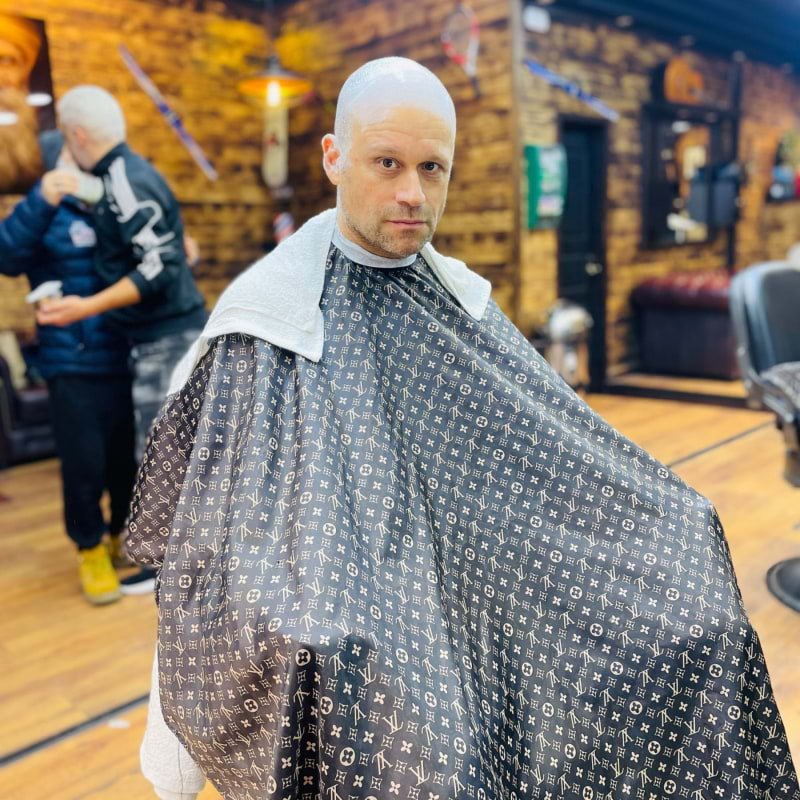 Man with a shaved head sitting in a barbershop chair, wearing a black cape with a white monogram pattern, and a white towel around his shoulders.
