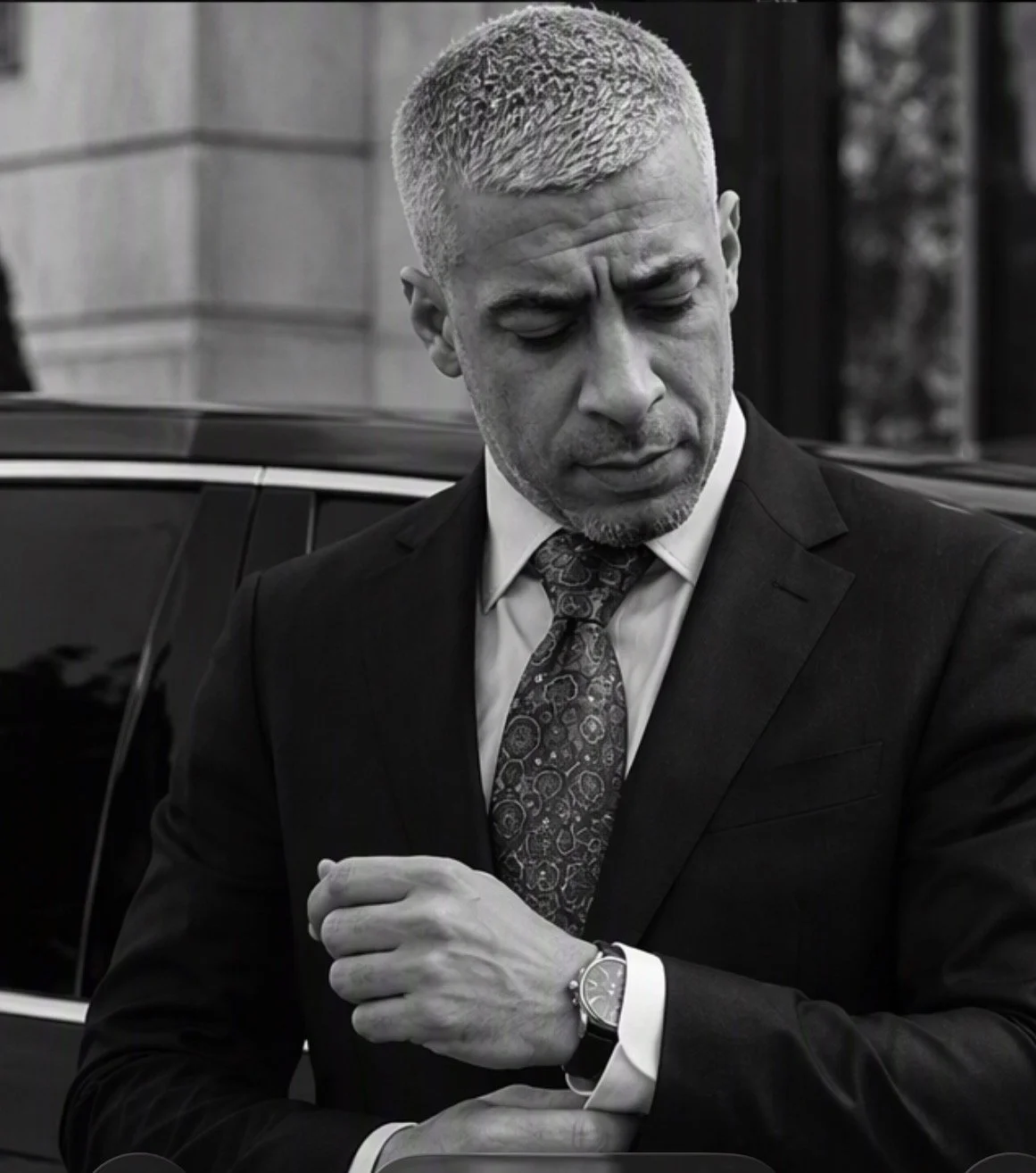 Black and white photo of a man in a suit adjusting his watch, with a serious expression, standing near a black car.
