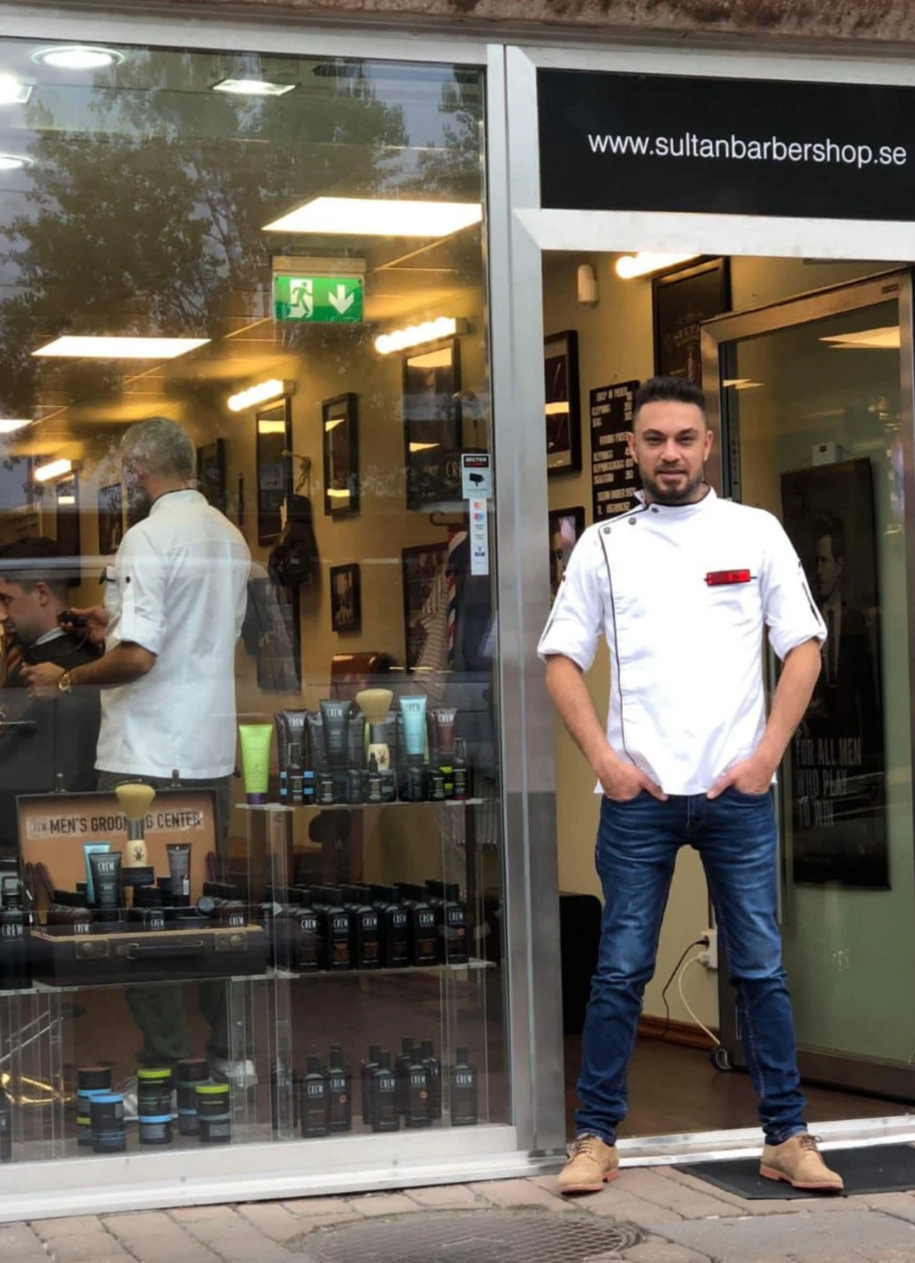 A young man standing outside a barbershop with a glass storefront. The man is wearing a white chef coat and jeans, with his hands in his pockets, looking at the camera. Inside the shop, a barber can be seen working on a customer. The shop displays grooming products and has a sign with the URL www.sultanbarbershop.se.