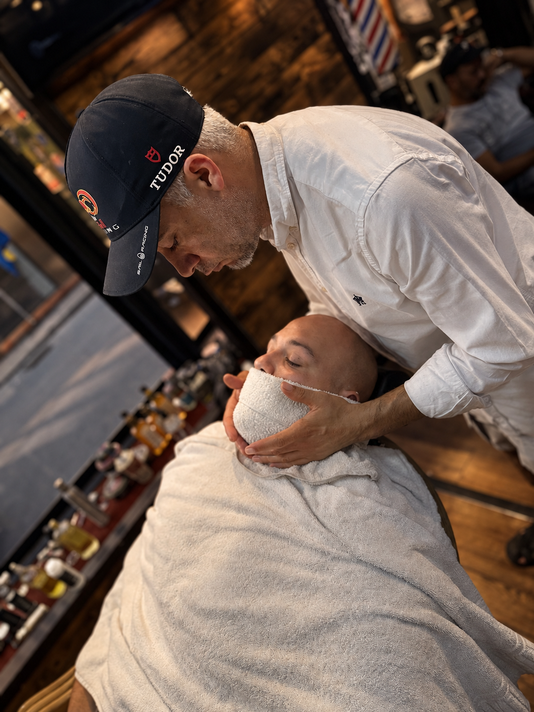 A barber performing a straight razor shave on a man with a shaved head in a barbershop. The barber wears a dark cap and white shirt, and is carefully shaving the man's face with a straight razor while the man has a towel wrapped around his neck. The barbershop has a wooden interior with various grooming products on the shelf.