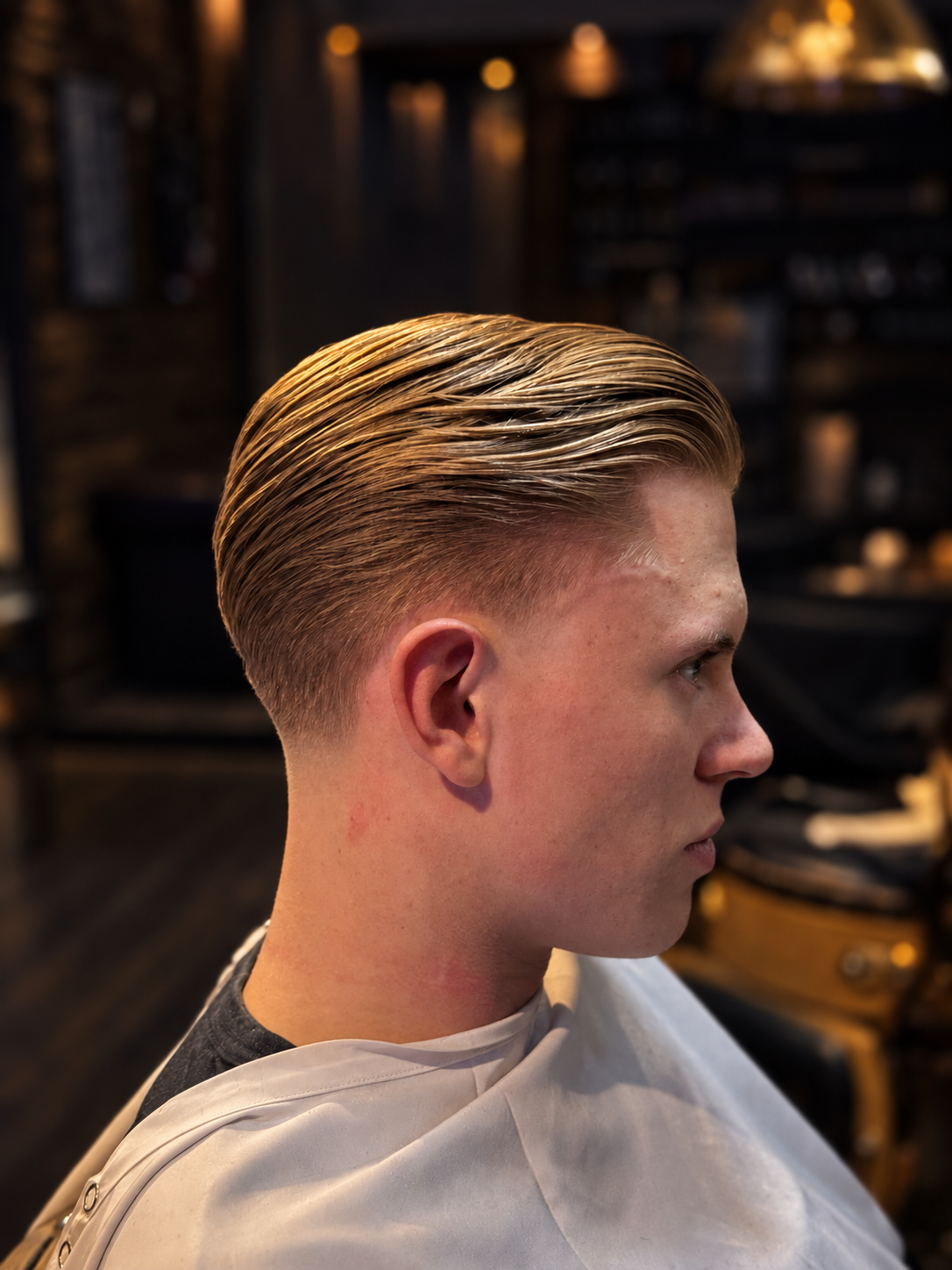 Young man with slicked back blonde hair sitting in a barbershop, wearing a white cape, with a blurred background of shelves and chairs.