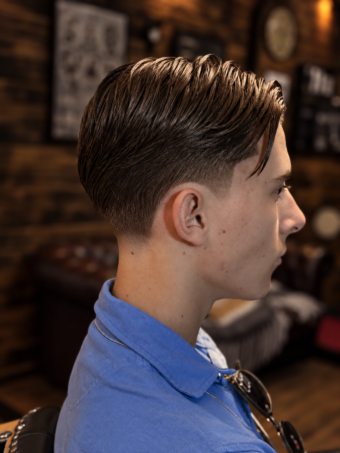 Side profile of a young man with a slicked-back hairstyle, wearing a blue collared shirt and sunglasses hanging from the collar, inside a barbershop.