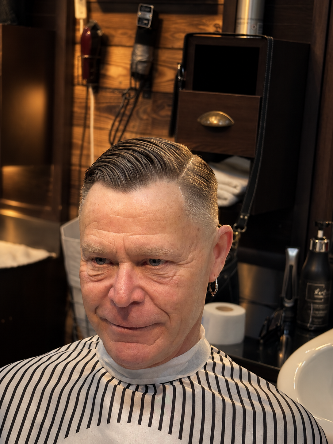 Man with slicked-back hair sitting in barber shop, wearing striped cape, surrounded by barber tools and products.