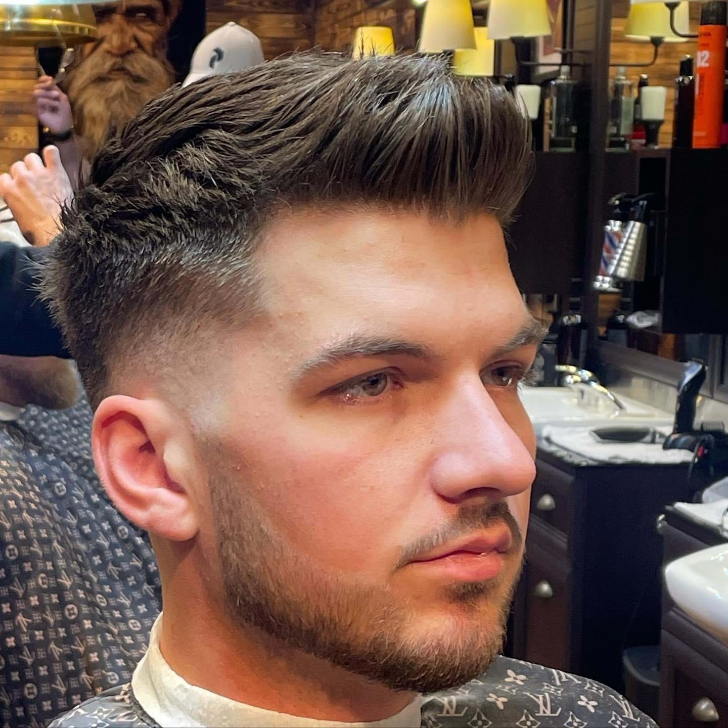 Close-up of a young man with a fresh haircut, sitting in a barbershop with barber equipment and shelves in the background.