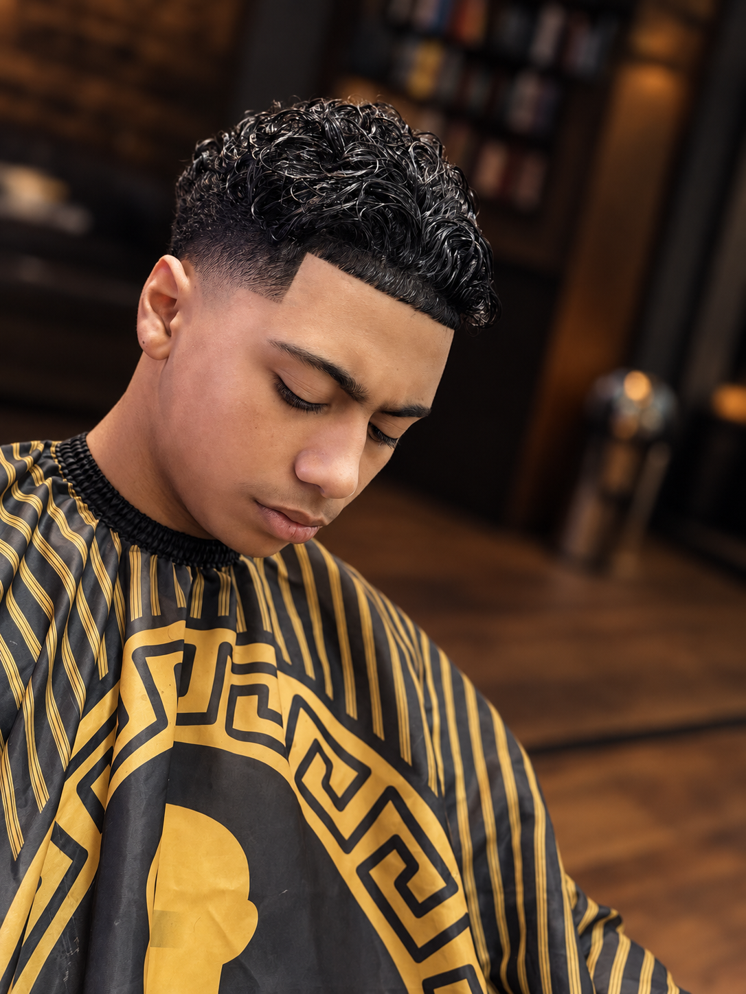 Young man with a modern crew haircut sitting in a barbershop, wearing a barber cape with gold and black designs, looking down with closed eyes.