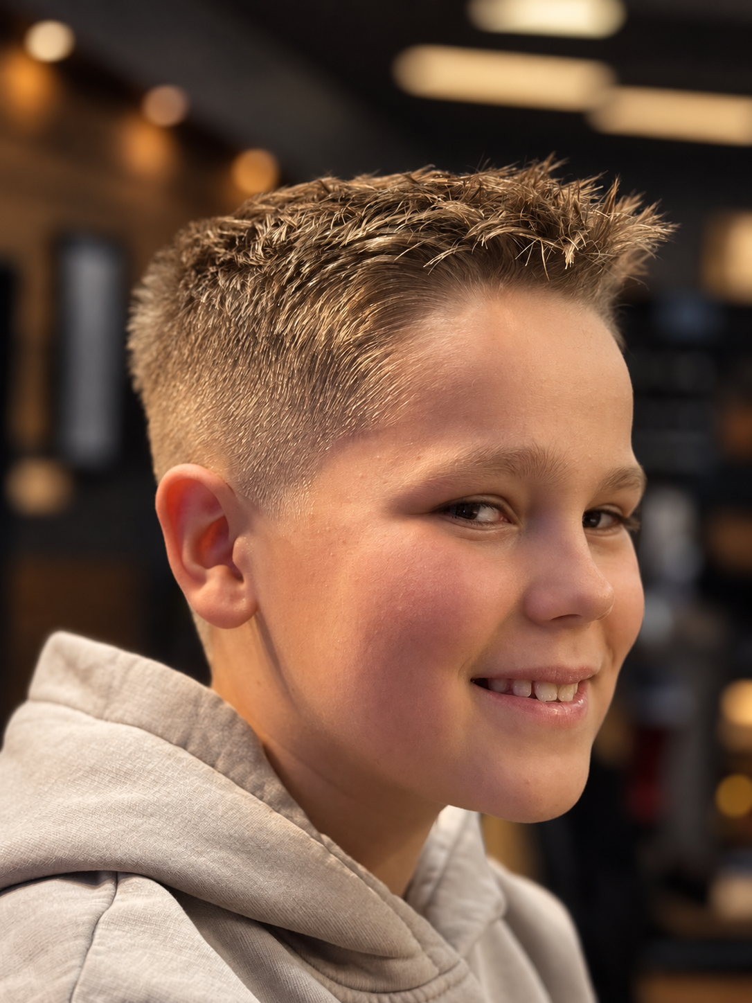 A young boy with a short, textured hairstyle smiling in a store or salon with blurred background.