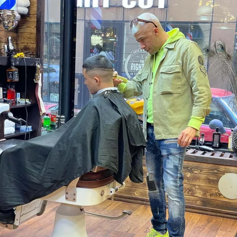 Barber giving a haircut to a young male client in a barbershop with wooden walls and large windows.