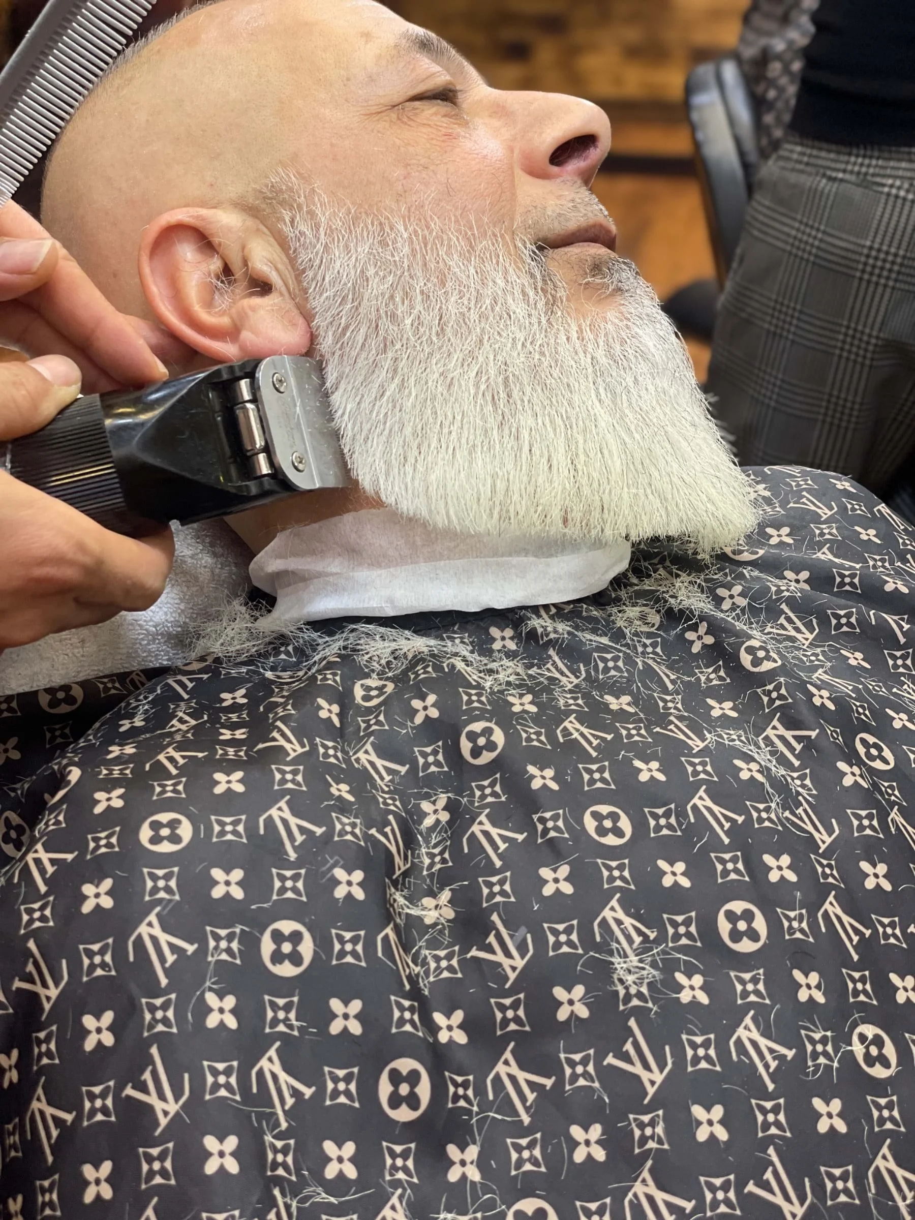 A man with a white beard receiving a haircut with an electric clipper at a barbershop.