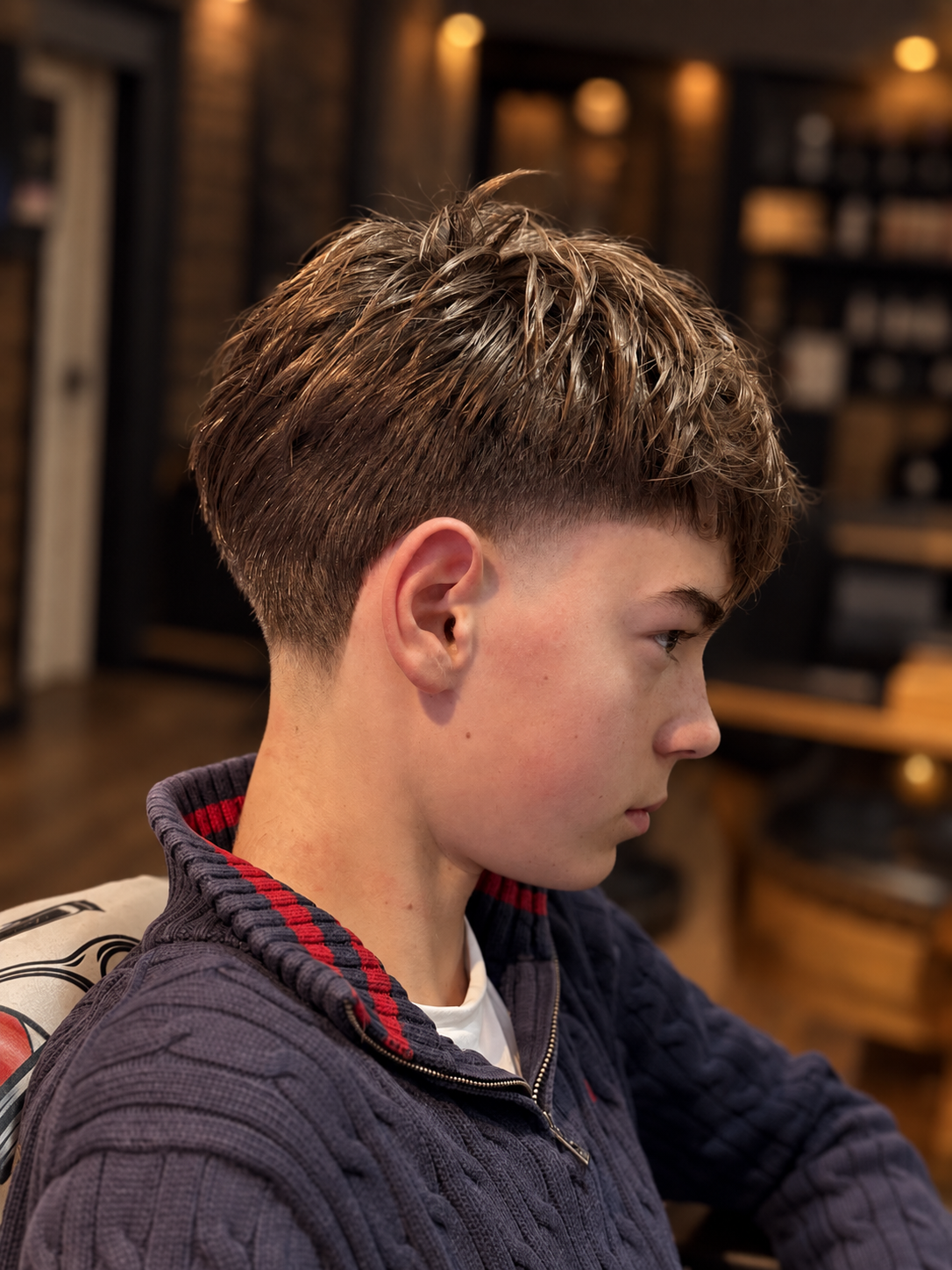 Profile of a teenage boy with a textured, two-tone haircut, wearing a dark jacket with red and white accents, sitting indoors with a blurred background of warm lighting and wooden furniture.