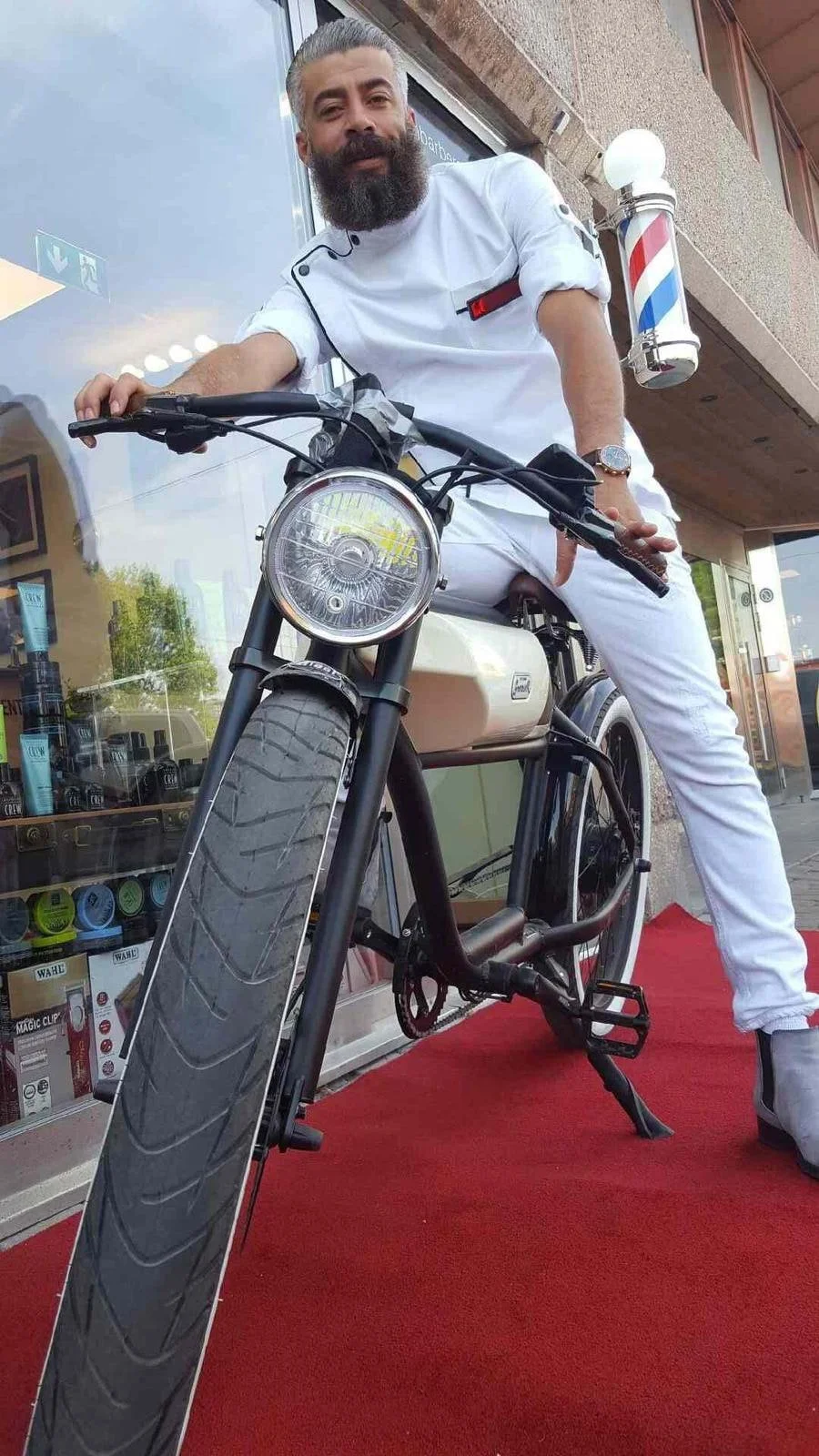 Man with beard and mustache wearing a white chef's jacket and white pants, sitting on a black electric bicycle with a large front tire, outside a shop with glass windows. The shop displays hair care products. The man is leaning forward slightly, resting his left hand on the handlebar, and looking at the camera. There is a barber pole mounted on the wall beside him. The scene is on a red carpet.