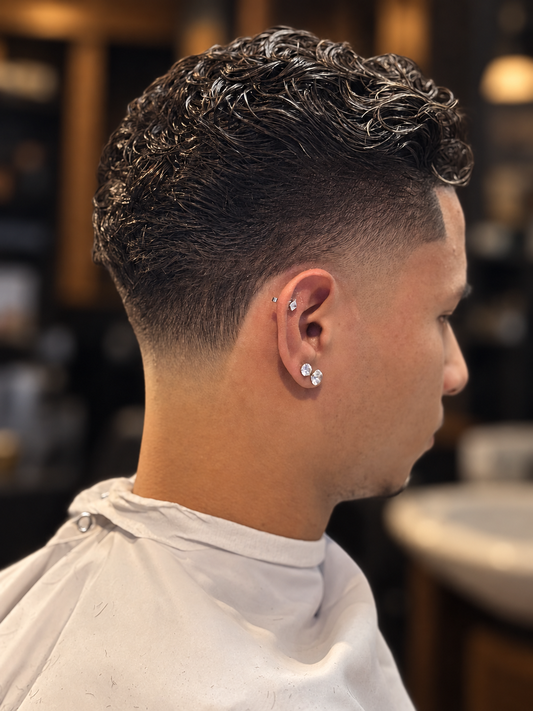 Close-up of a person with a short, faded haircut and curly styled hair on top, wearing multiple earrings, in a barber shop setting.
