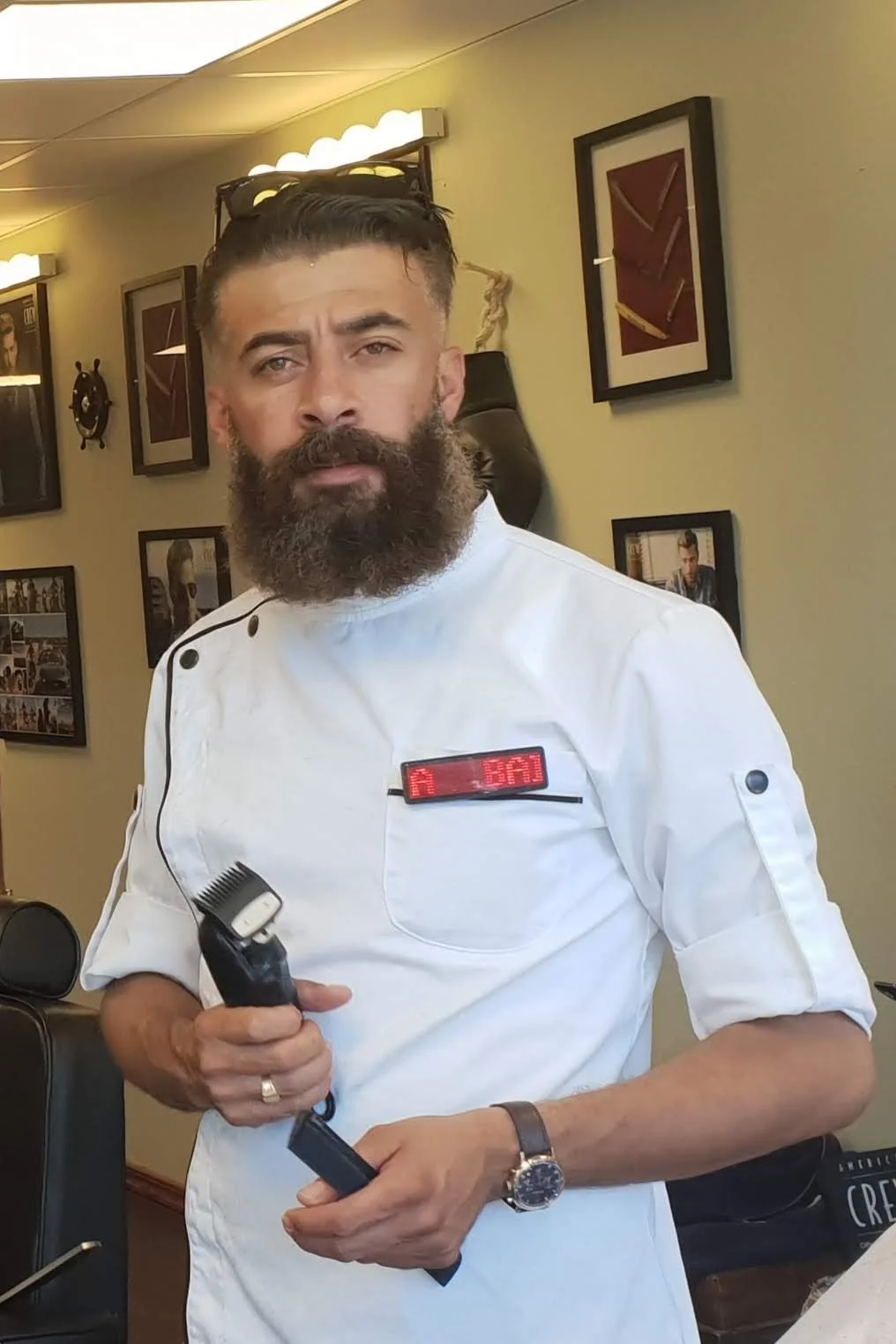 A bearded man wearing a white chef's coat with glasses on his head, holding a barber clipper, standing in a room with framed pictures on the wall.