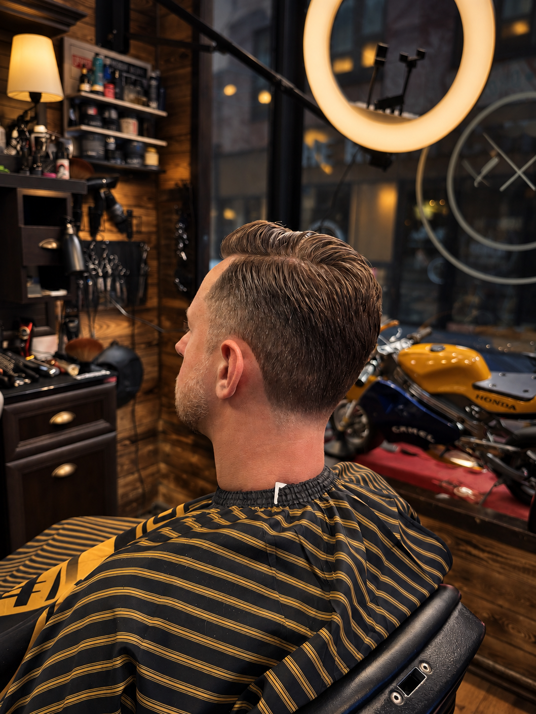 Man with styled hair sitting in barber chair at barbershop, with motorcycle and tools visible in the background.